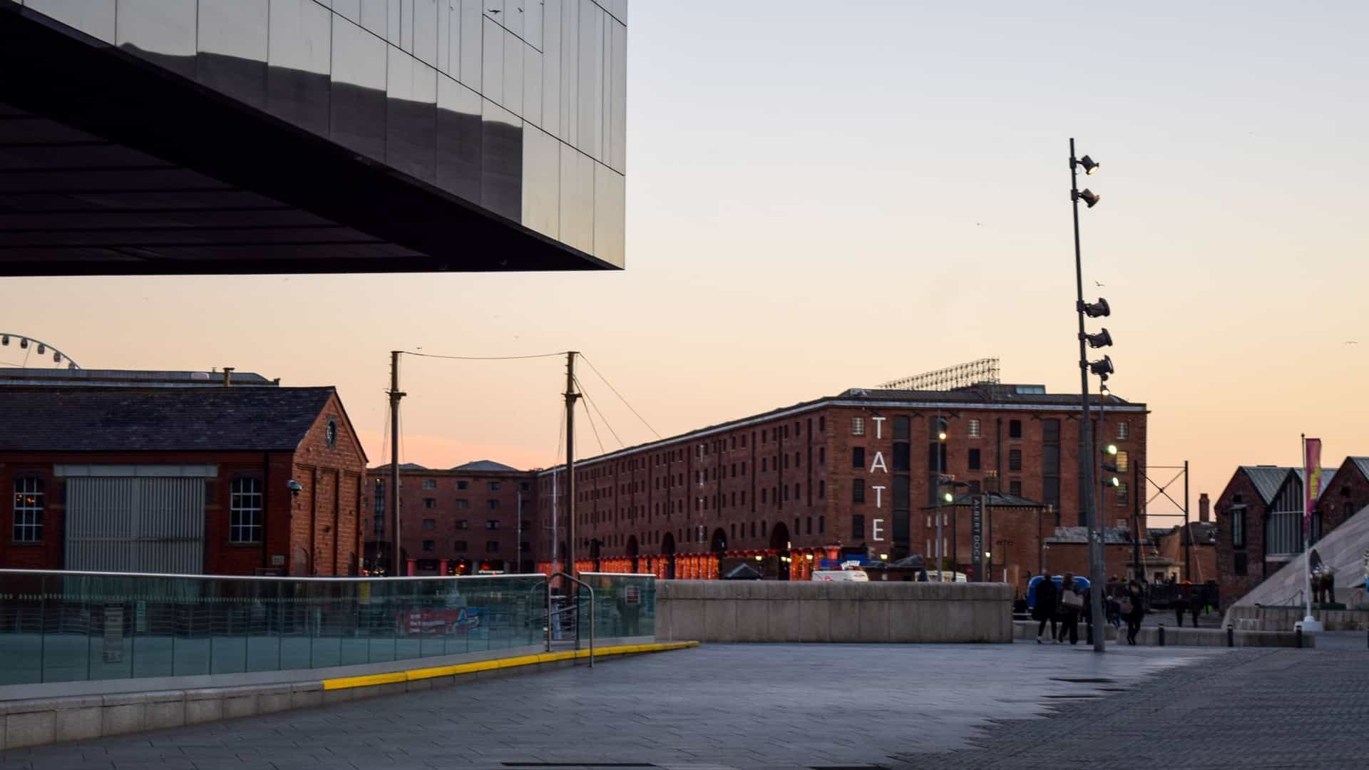 A dusk view of the Liverpool waterfront with a paved open plaza in the foreground. Dominating the background is a long, historic red-brick warehouse (part of the Royal Albert Dock), with the large, white block letters spelling "TATE" visible on its facade. To the left, a corner of a reflective, modern black or dark glass building overhangs the scene. The sky is a soft orange and pink from the sunset, and a few people are walking near a set of dockside railings.