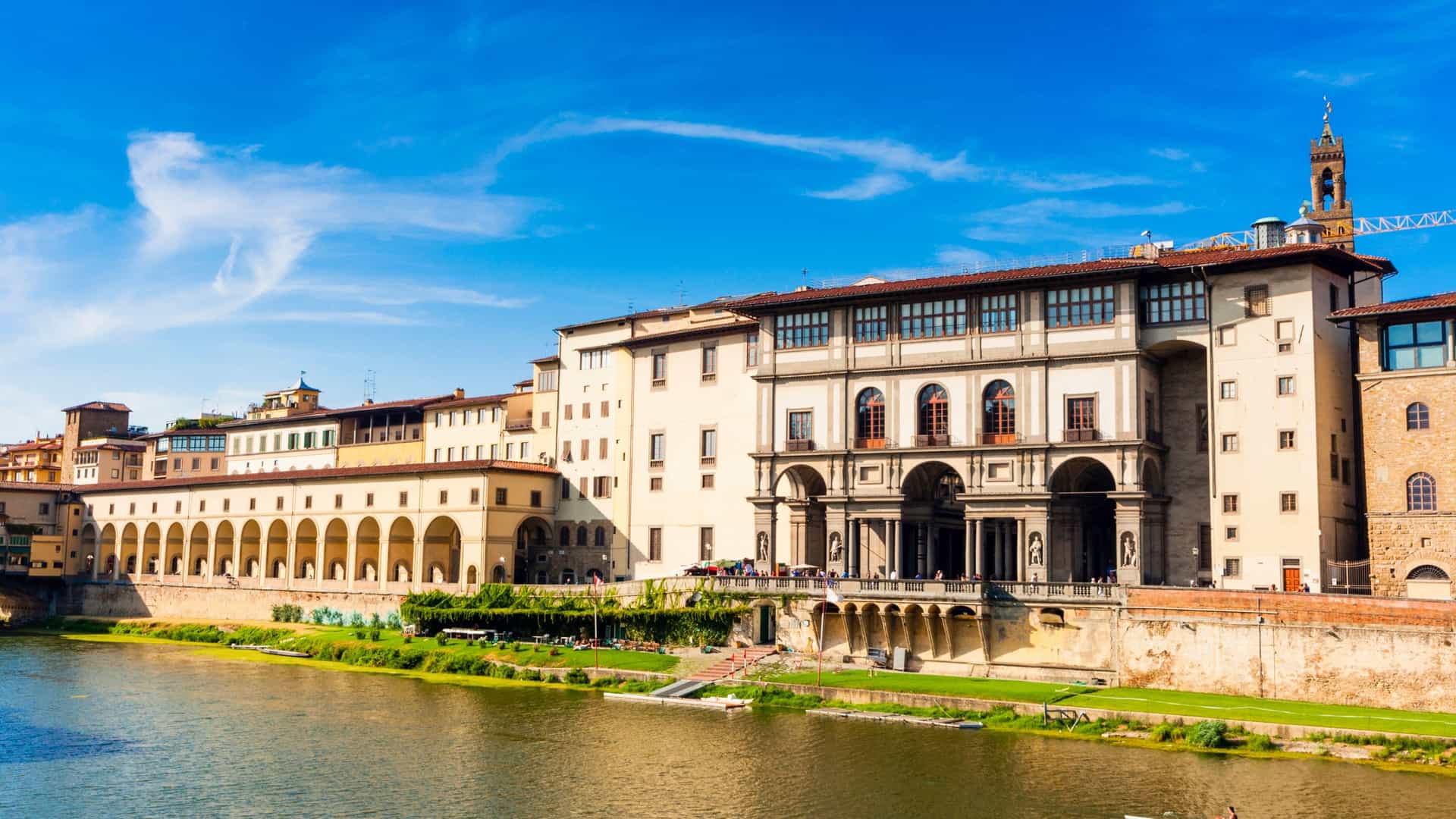 A classic view of the Uffizi Gallery along the Arno River in Florence, Italy, part of the city's UNESCO World Heritage site, showing the distinctive U-shaped Renaissance palace architecture with a statue visible in the foreground.