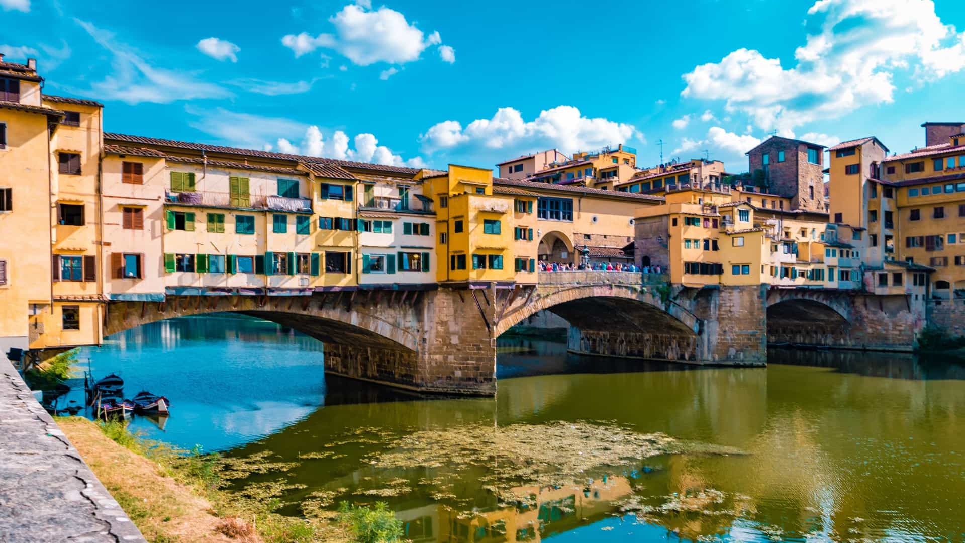 A scenic, daytime view of the Ponte Vecchio bridge spanning the Arno River in Florence, Italy, featuring the iconic medieval shops built directly along its length.