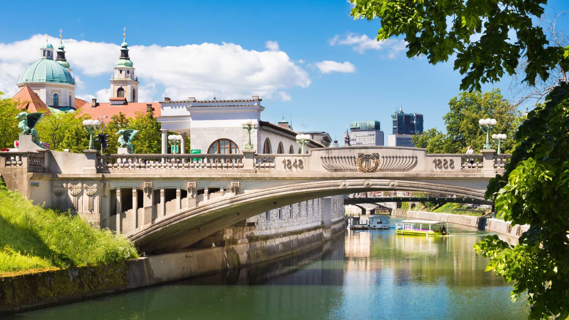 A cityscape of Ljubljana, Slovenia, featuring the Dragon Bridge (Zmajski most) spanning the Ljubljanica River. The Art Nouveau style bridge is a technical monument known for its four famous sheet copper dragon statues, which are a symbol of the city.