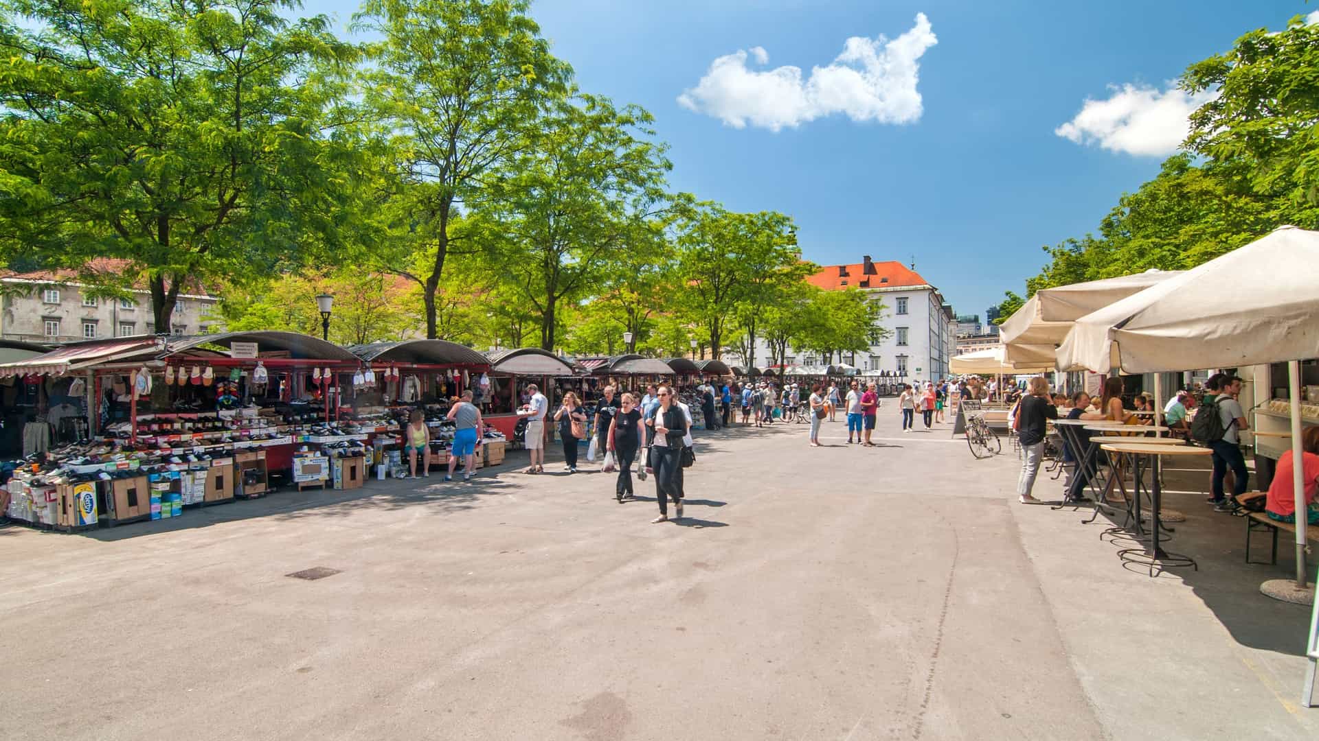 People walking and shopping in a central market in Ljubljana, Slovenia, on a bright, sunny day. The image shows the open air market with stalls and crowds, with the prominent green domes and towers of St. Nicholas Church in the background.