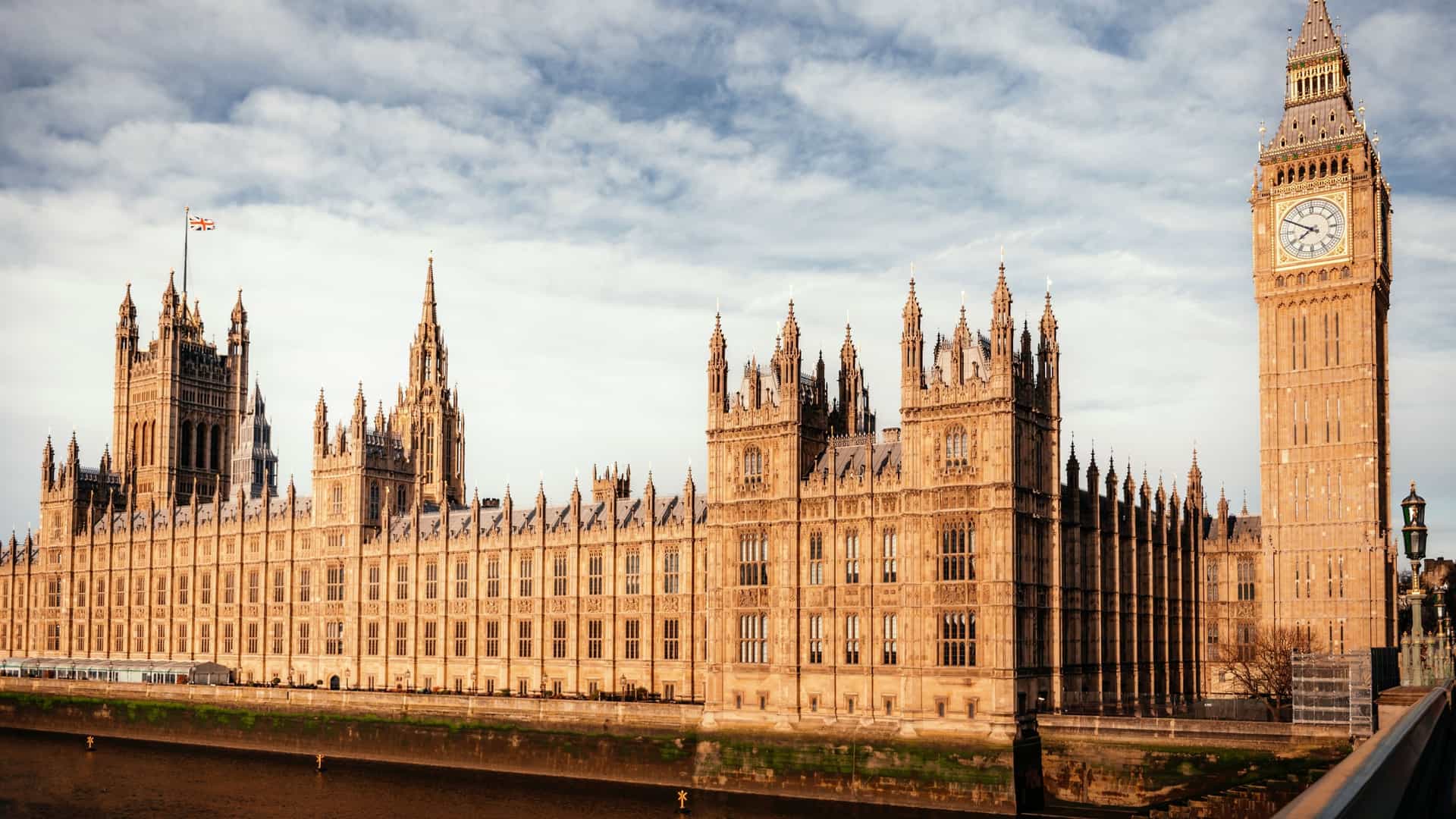 A view of the iconic Houses of Parliament and the Elizabeth Tower, known as Big Ben, from across the Thames River, with Westminster Bridge in the foreground. The Gothic Revival architecture of the building is visible under a blue sky with some clouds.