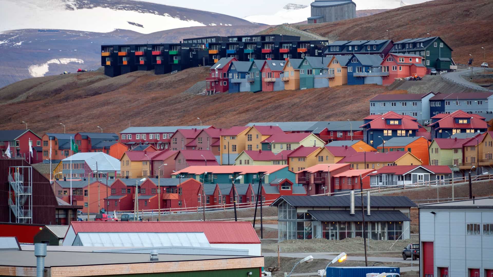 A scenic view of the colorful houses and buildings of Longyearbyen, the largest settlement in Svalbard, nestled in a valley. The town is surrounded by large, steep mountains partially covered in snow and a fjord in the distance.