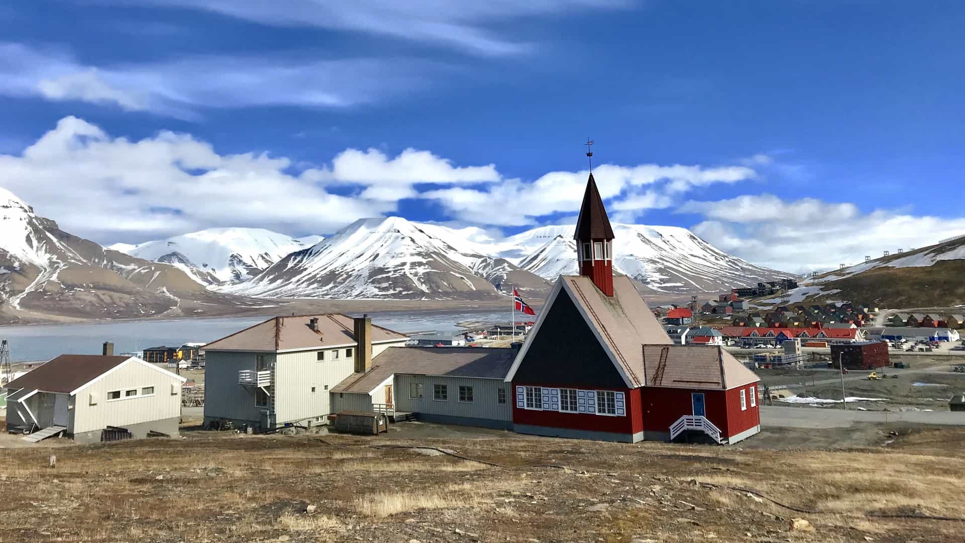 The Svalbard Church, the world's northernmost church, stands in the foreground of Longyearbyen town, featuring a light-colored wooden structure with a red roof. The building is surrounded by small residential homes and is set against a stunning Arctic mountainscape under a clear sky.