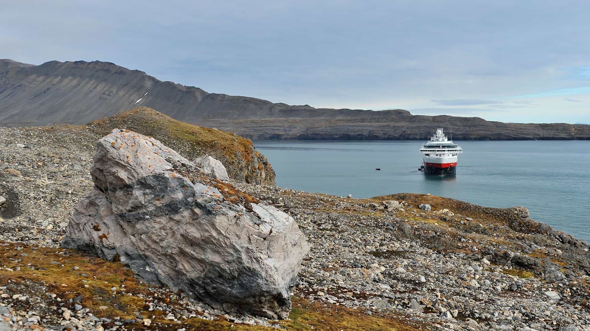 A large cruise ship sails through a fjord in the Svalbard archipelago. Towering, snow covered mountains and glaciers line the waters, with chunks of ice floating nearby. The sky is bright and clear, reflecting off the calm water.