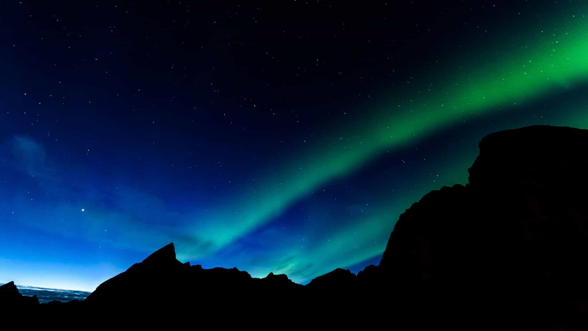 A brilliant green and pink Aurora Borealis (Northern Lights) dances across the dark night sky over snow-covered mountains, casting a perfect reflection on the still water of a fjord in Svalbard, Norway. The long exposure captures the ethereal glow of the light show.