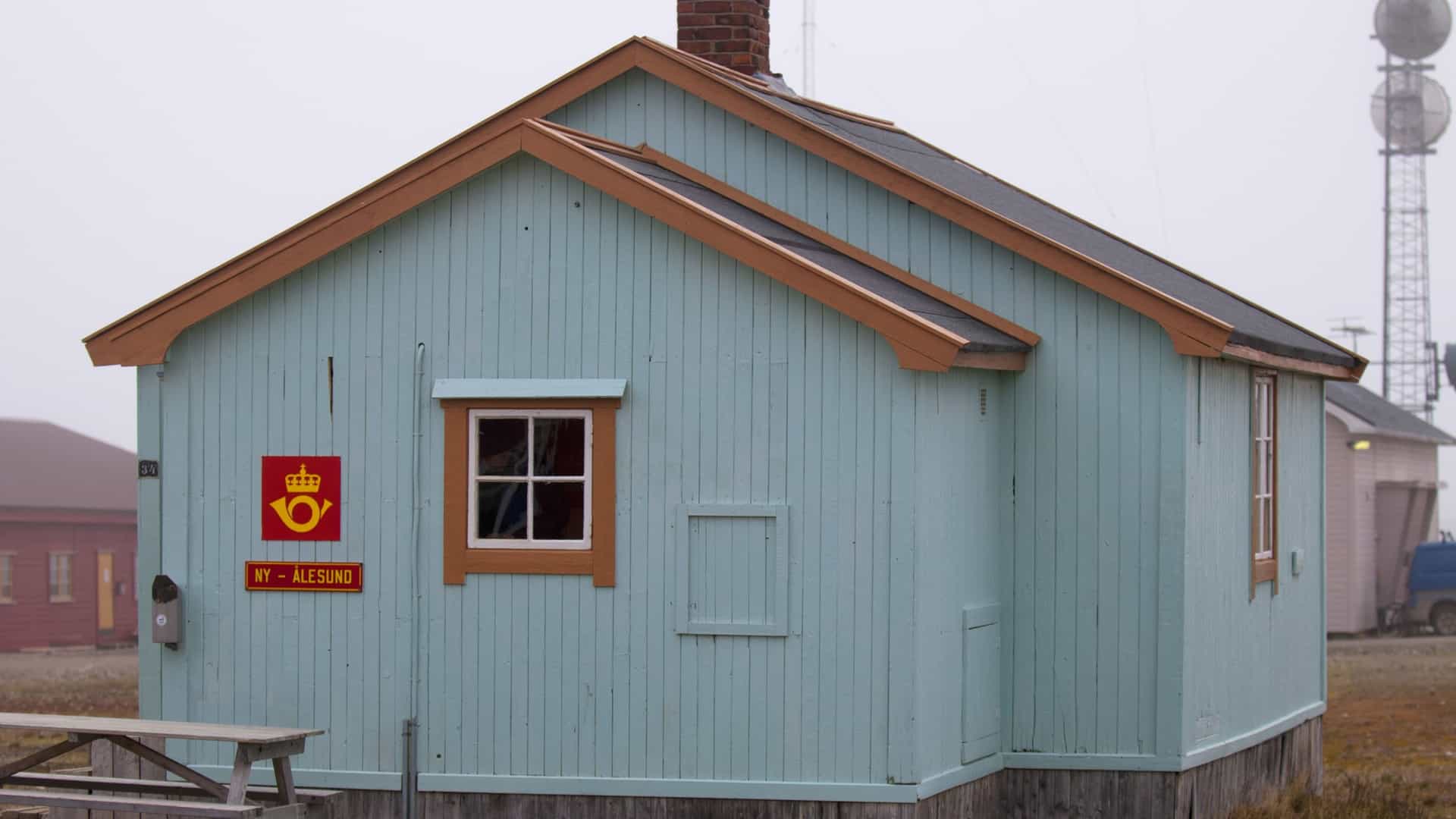 A photograph of the Svalbard Post Office in theArctic settlement of Ny-Ålesund, often cited as the northernmost post office in the world. The image shows the characteristic, small, brightly colored wooden building against a backdrop of snowy mountains and a cold, clear Arctic environment.
