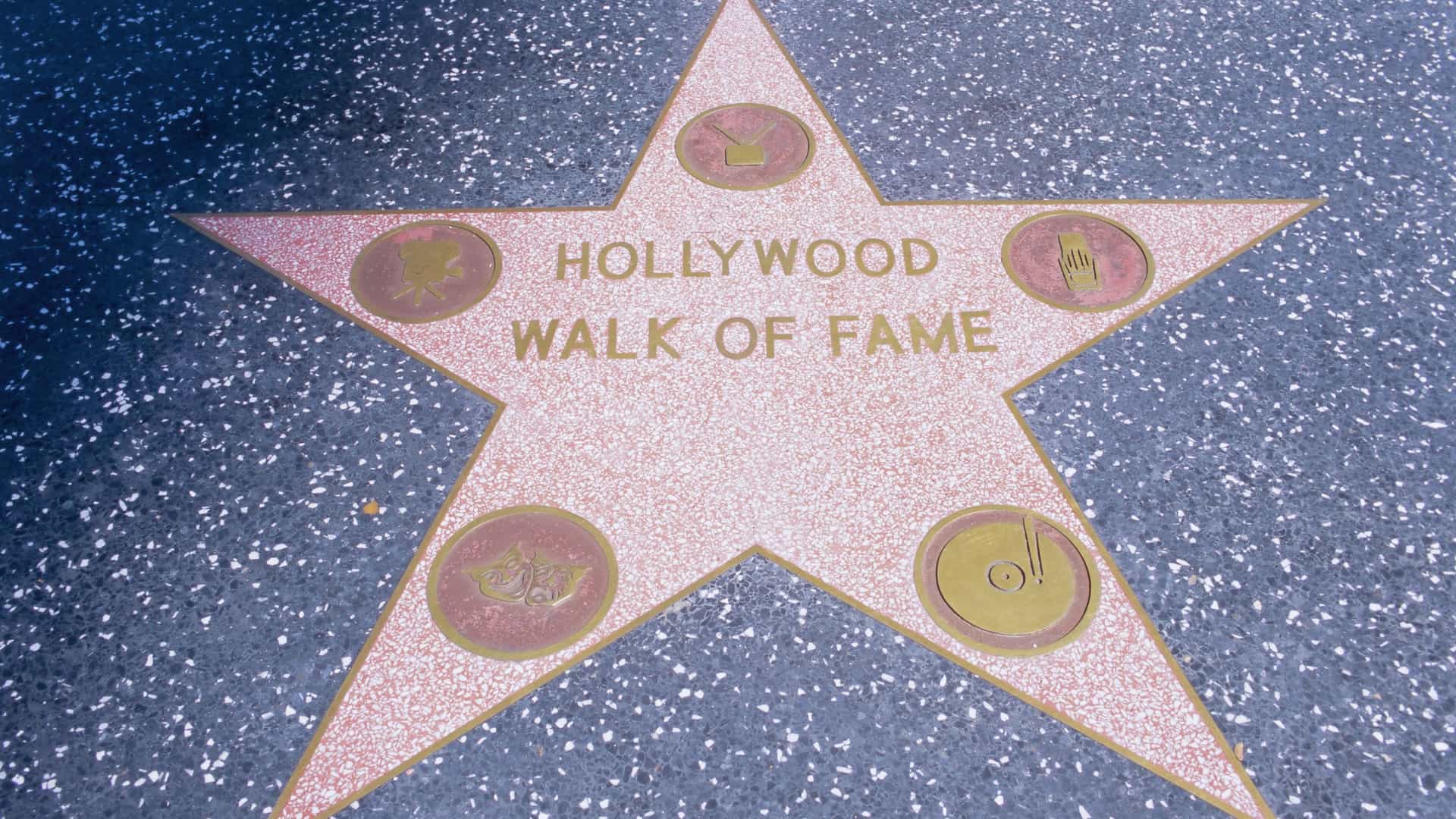 A close-up view of the Hollywood Walk of Fame on Hollywood Boulevard in Los Angeles, showing a five-pointed, coral-pink terrazzo star with a brass border and an engraved name and emblem. The star is embedded in the charcoal-colored sidewalk, with the busy street and surrounding buildings visible in the background.
