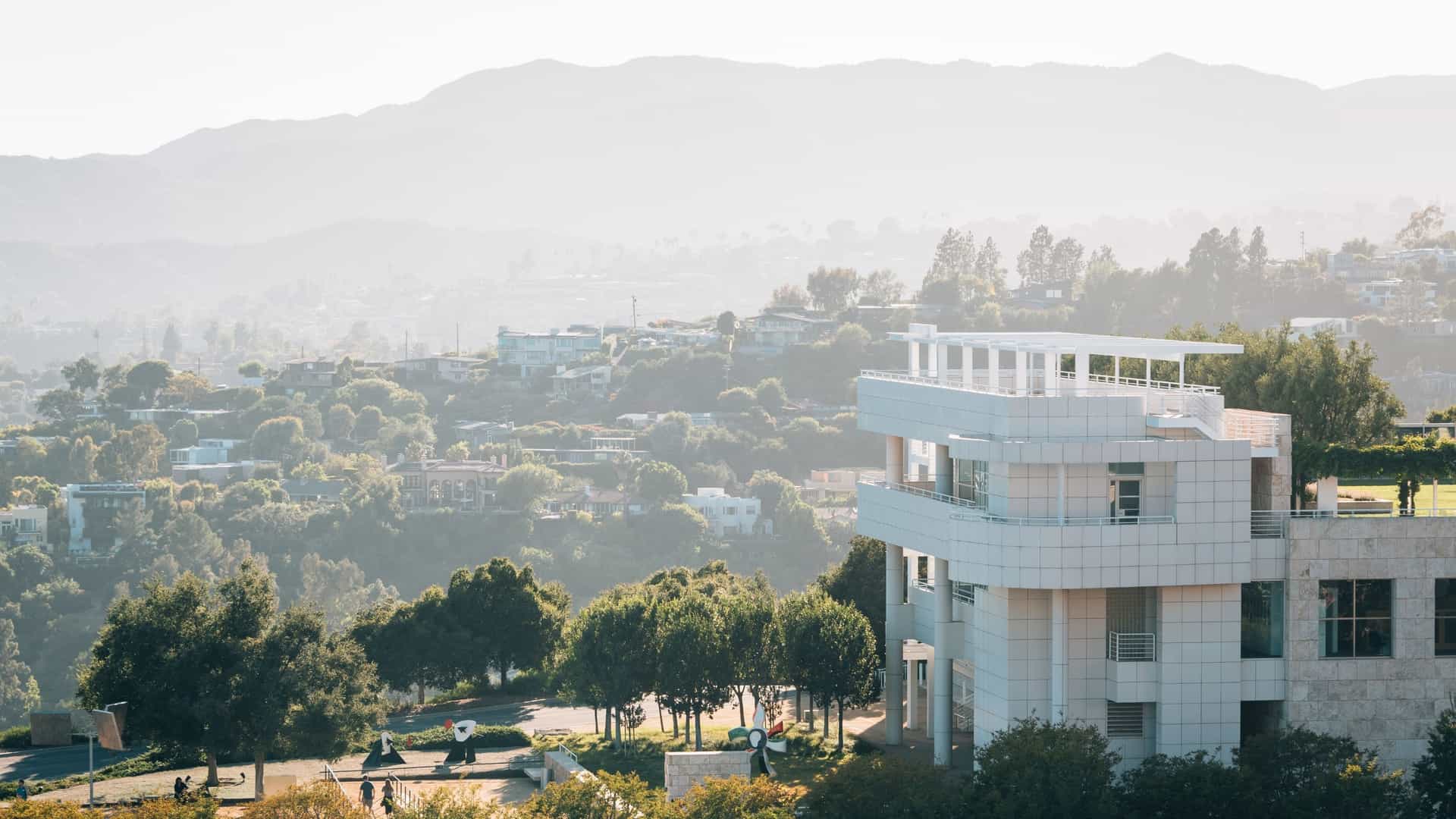 A panoramic view from a terrace at the Getty Center in Los Angeles. The foreground shows the modern, light-colored travertine and glass architecture of the museum, with the vast, hazy Santa Monica Mountains stretching into the distance under a bright sky.