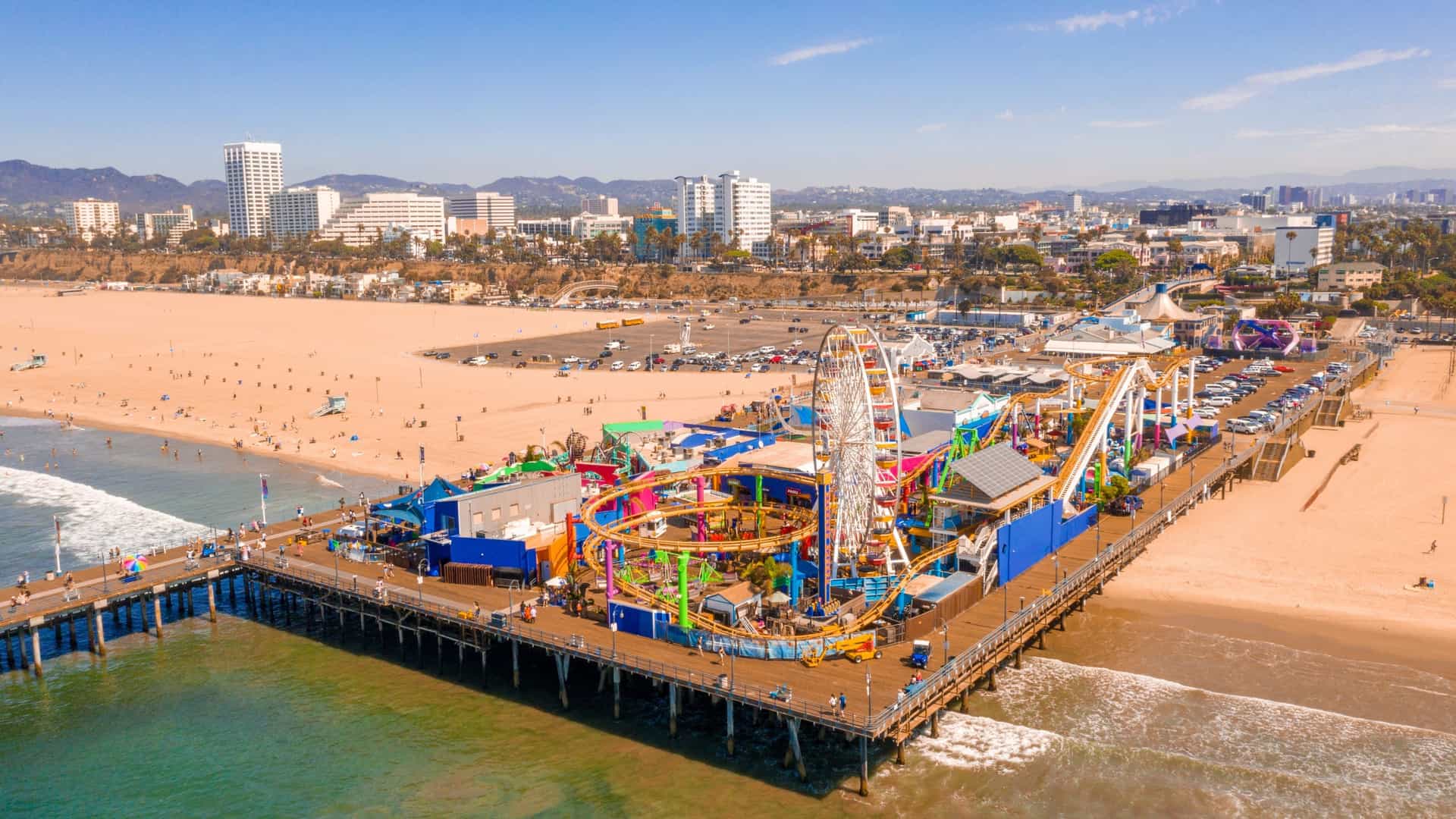 An aerial view of the Santa Monica Pier in California. The pier juts out into the Pacific Ocean, featuring an amusement park with a Ferris wheel and roller coaster. The long sandy beach is visible to the sides, with the city of Santa Monica in the background. The ocean has waves rolling towards the shore.
