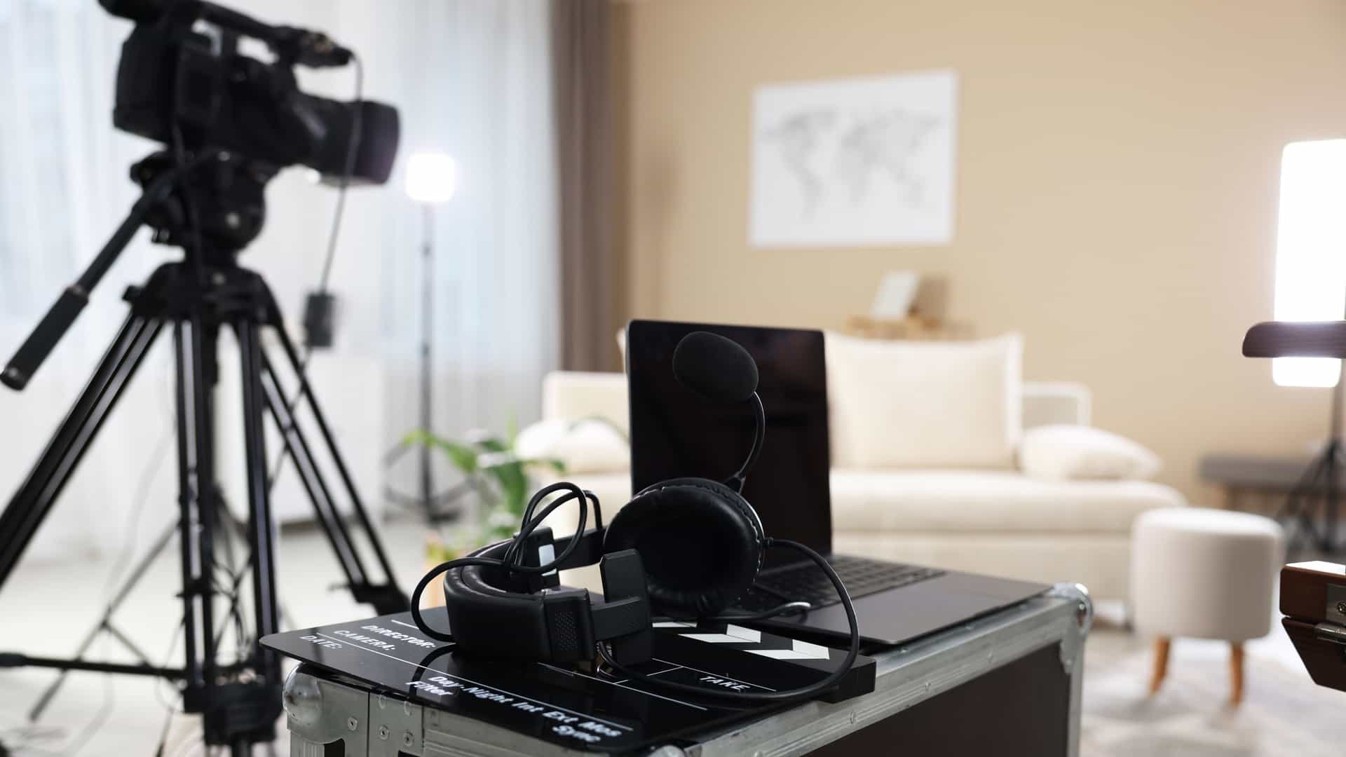 A professional video camera on a tripod is set up in a living room, suggesting a film production or broadcast. In the foreground, a laptop, headphones, and a microphone are arranged on a flight case. The scene evokes a behind-the-scenes look at a modern Hollywood Studios setup for content creation.