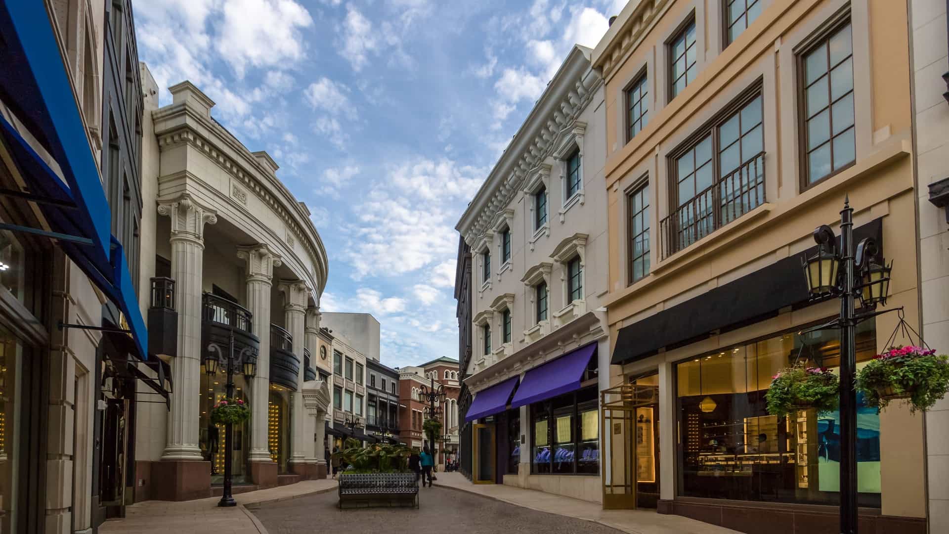 A bright, sunlit view down Rodeo Drive in Beverly Hills, California, showing a street lined with palm trees in the median and luxurious high-end designer stores and boutiques with ornate window displays.