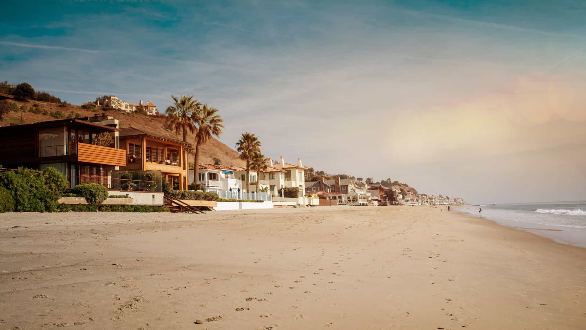 A serene beach in Malibu, California, at sunset. The sky is painted with warm orange, pink, and purple hues as the sun sets over the Pacific Ocean. The foreground features a sandy beach and gentle waves, with large, dramatic rock formations visible in the distance. The setting sun reflects off the wet sand, creating a golden glow.