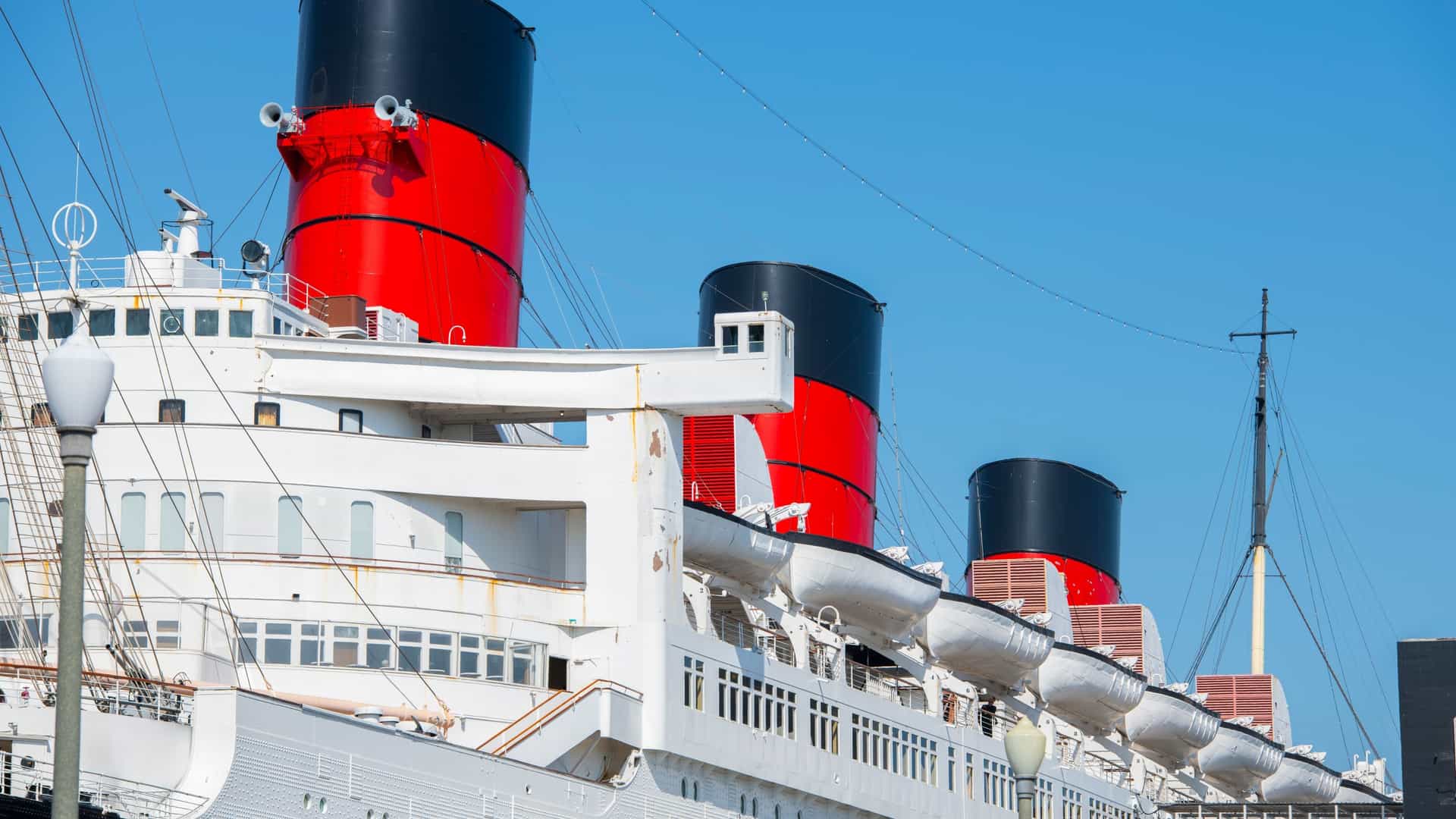A large, vintage ocean liner, the RMS Queen Mary, is permanently docked in the harbor of Long Beach, California. The ship is lit up against a night sky, with the city skyline and harbor lights visible in the background. The water in the foreground reflects the lights.