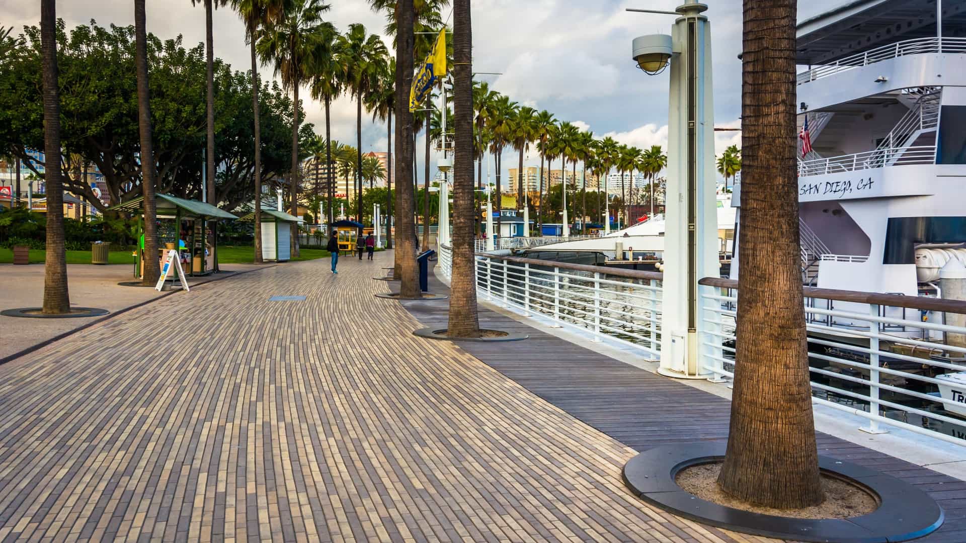 A scenic waterfront walkway in Long Beach, California, with palm trees and modern buildings lining the path. The walkway overlooks a marina with boats docked in the water, and the downtown skyline is visible in the background under a sunny sky.