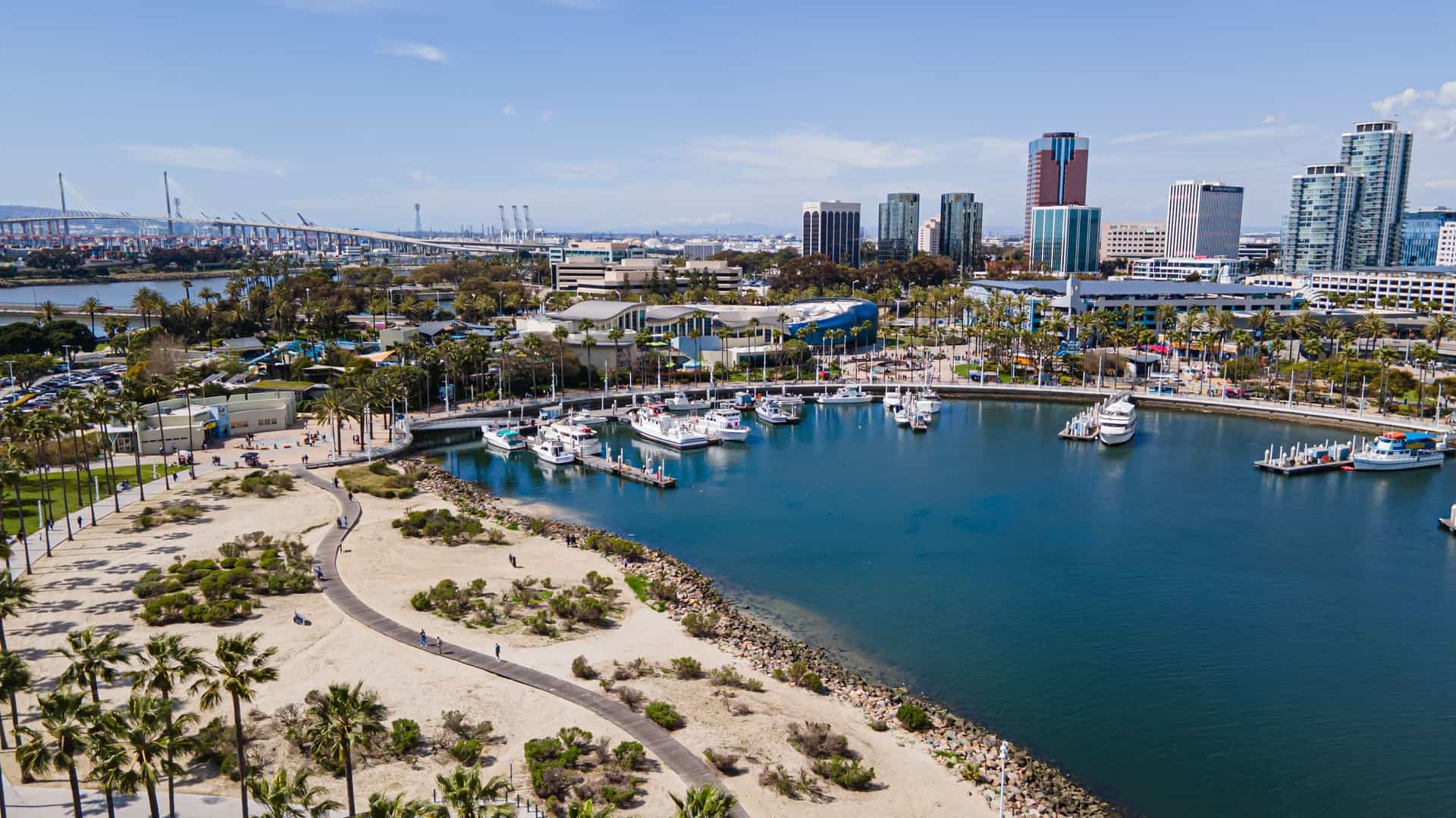 A scenic view of Shoreline Village in Long Beach, California, an entertainment and dining center. The image features the area's distinct, brightly colored, New England-style buildings clustered along the waterfront, overlooking the Rainbow Harbor filled with boats, with the Long Beach city skyline or the Queen Mary often visible in the background.