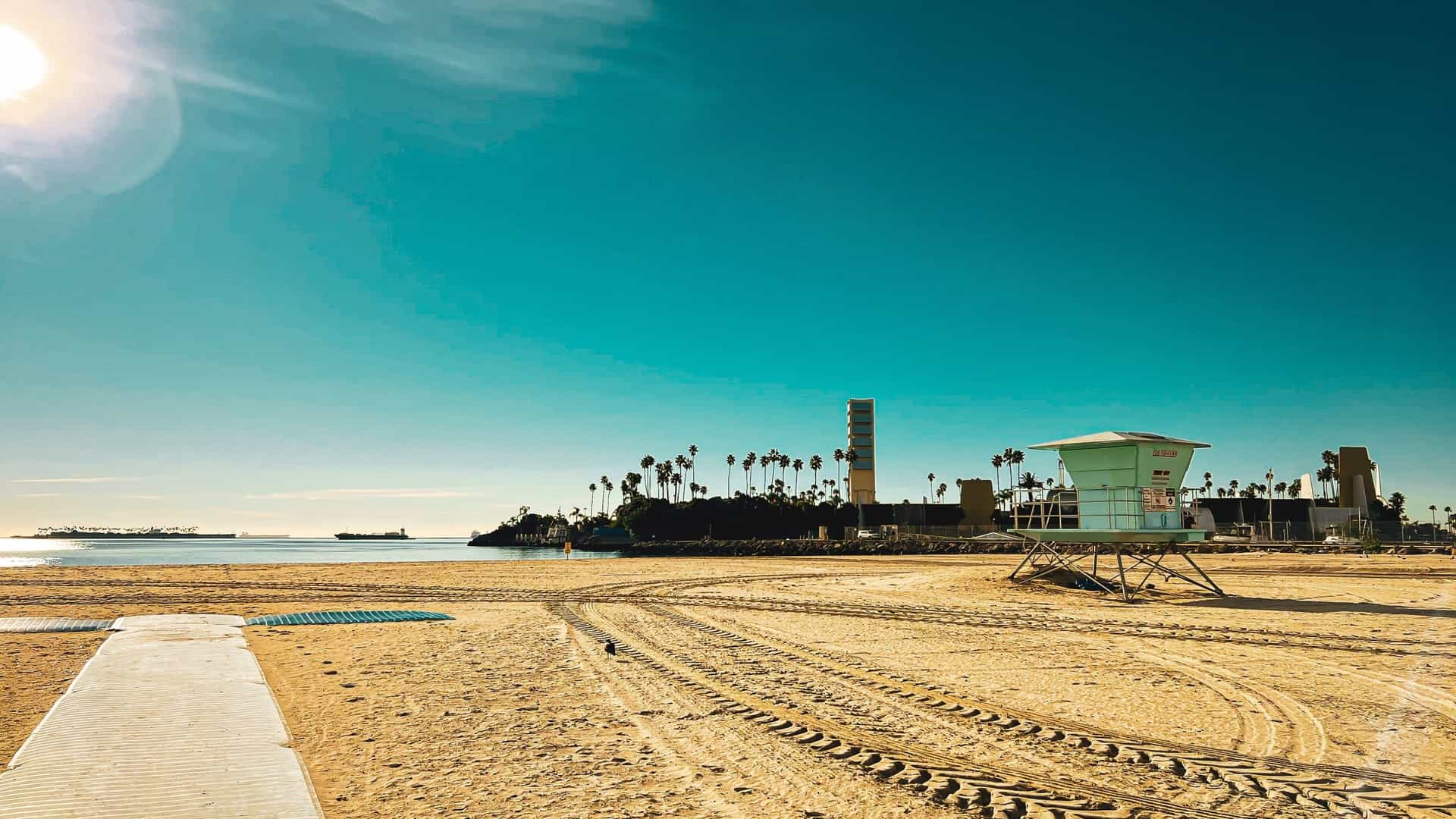 A stunning photo of Alamitos Beach in Long Beach, California, captured during the golden hour sunset. The image features golden sand leading to the sparkling ocean, with a classic lifeguard tower visible on the shoreline, highlighting the vibrant and popular coastal atmosphere.