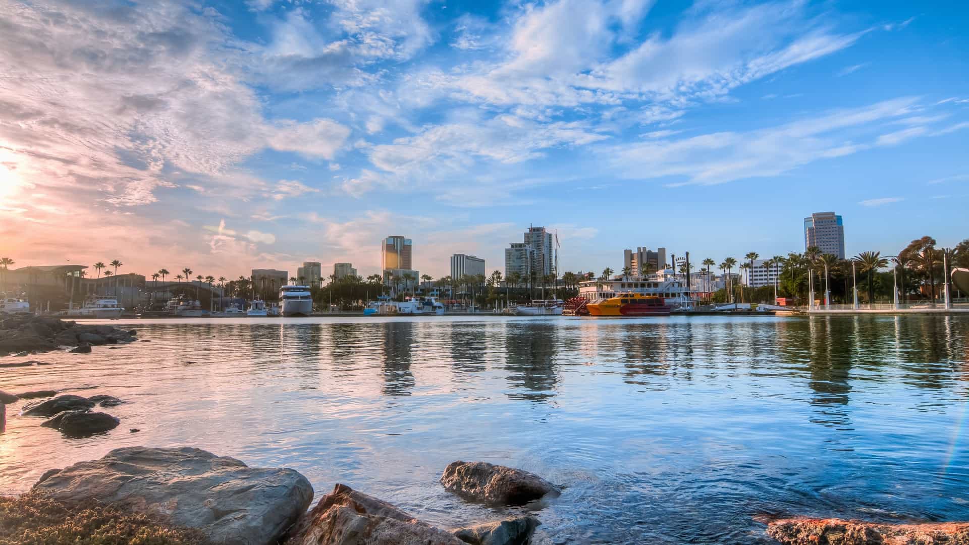 A sunny, wide-angle view of the Long Beach Harbor and marina area. The image features rows of docked yachts and smaller boats in the foreground, with the modern, distinctive buildings of the downtown skyline rising in the background. Prominent landmarks visible include the Aquarium of the Pacific complex and nearby hotels.