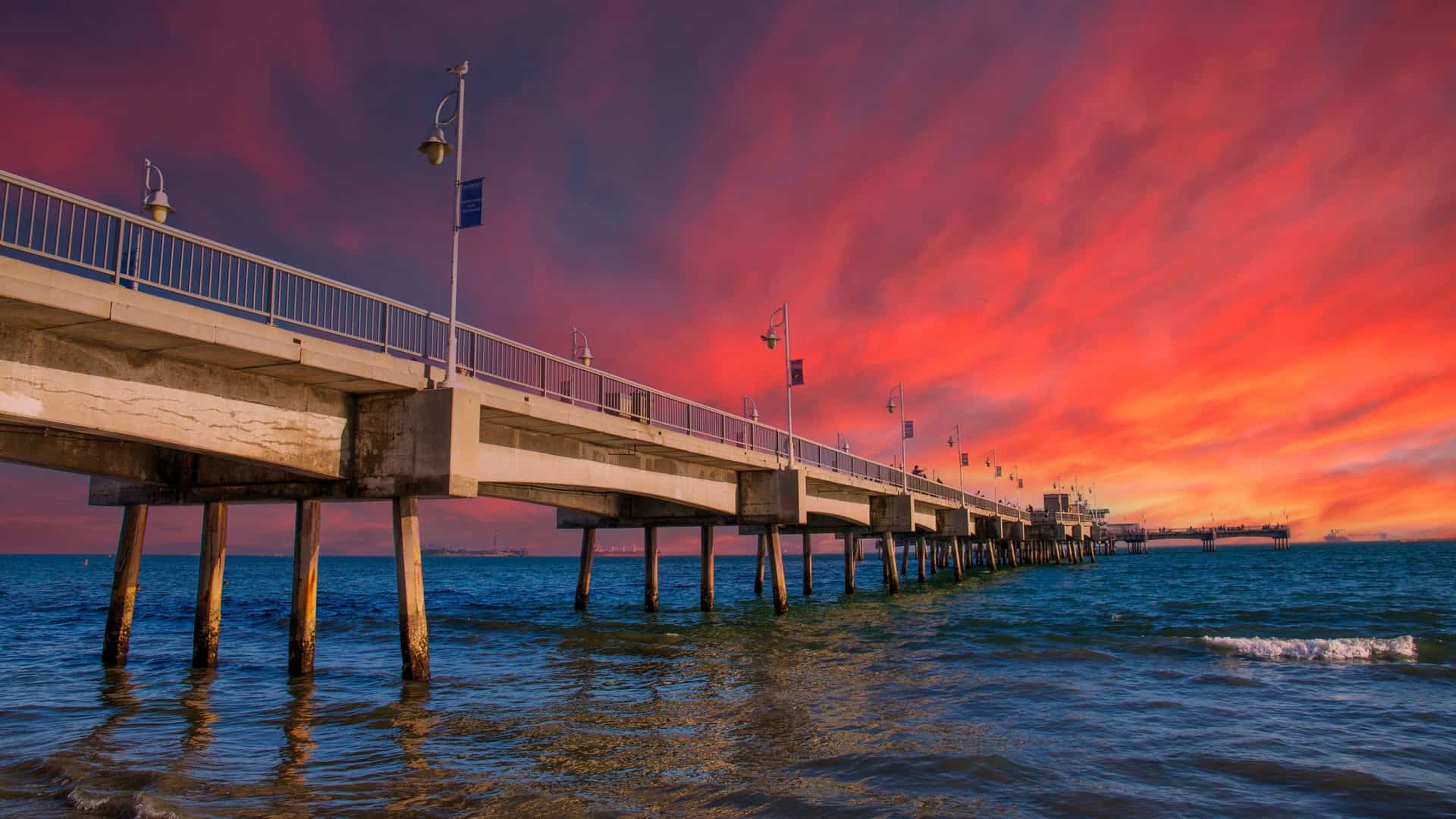A low-angle view of the Belmont Veterans Memorial Pier in Long Beach, California, extending out over the dark blue ocean water. The pier is dramatically backlit by an intense, vibrant sunset with a sky dominated by fiery red, orange, and deep purple clouds.
