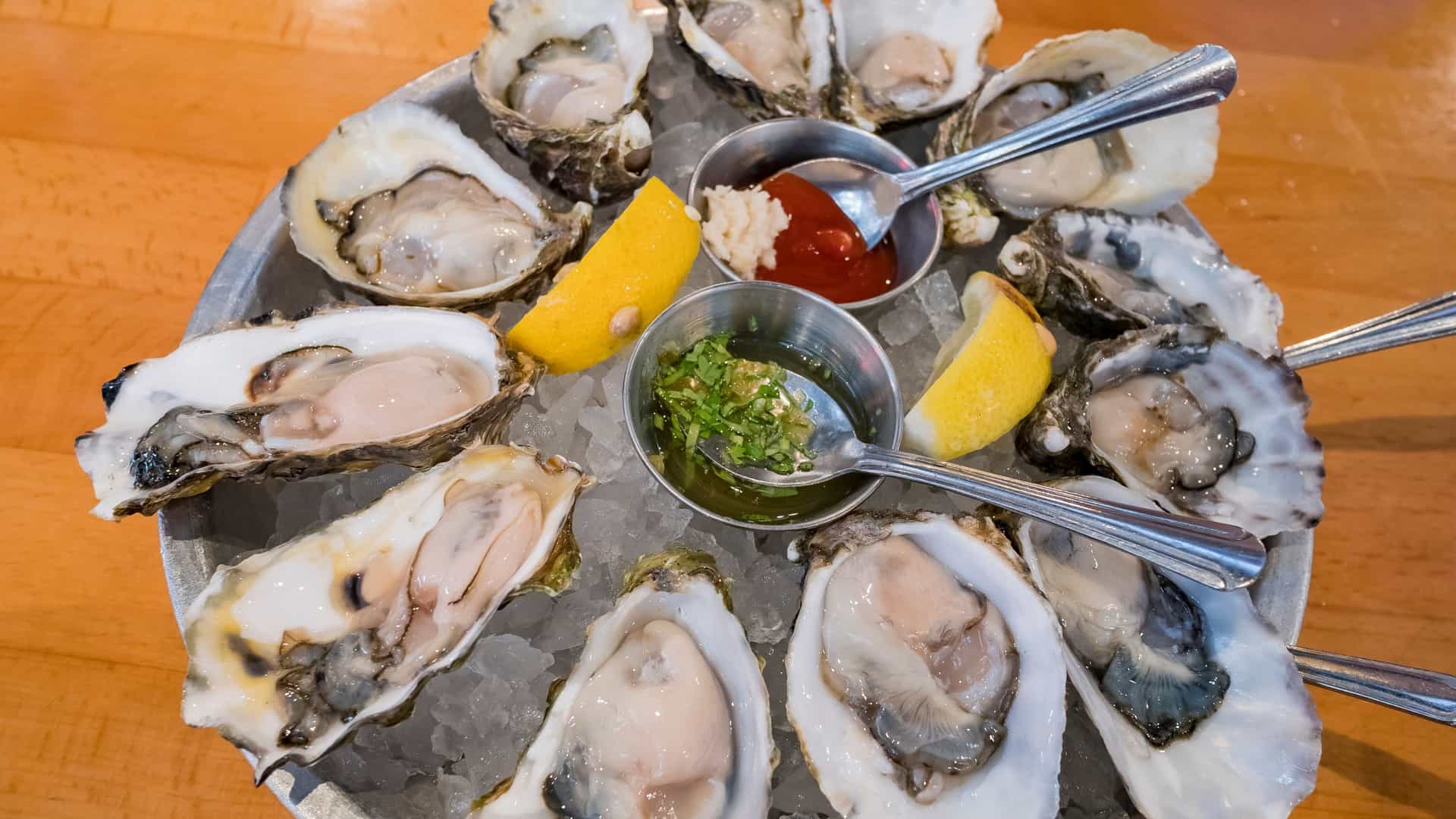 A close-up shot of a single raw oyster on the half shell resting on a white plate, garnished with a wedge of lemon. The image highlights the glistening texture of the fresh oyster meat, suggesting a gourmet seafood appetizer.