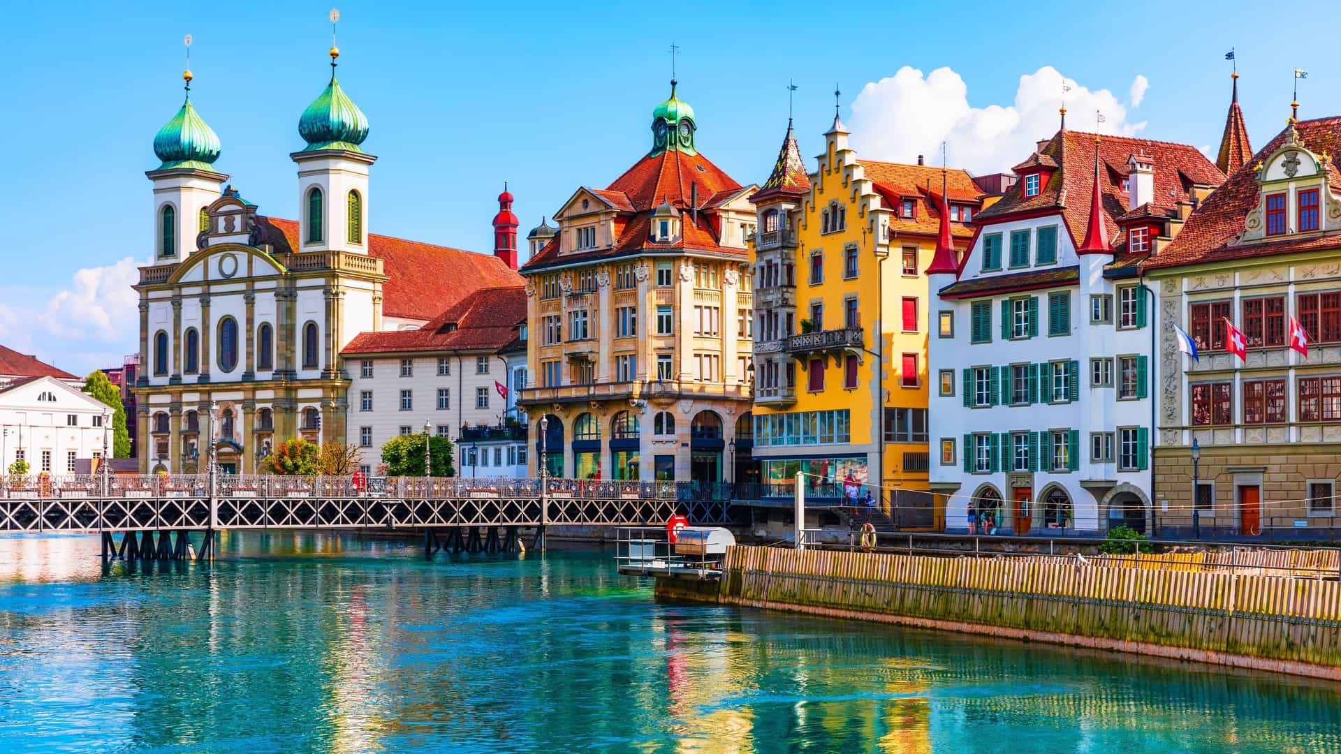 A scenic aerial panorama of the Old Town of Lucerne, Switzerland, in the summer. The image captures the medieval architecture, including rooftops, the Reuss River, and the surrounding landscape, providing a broad view of the city's historic center from above.