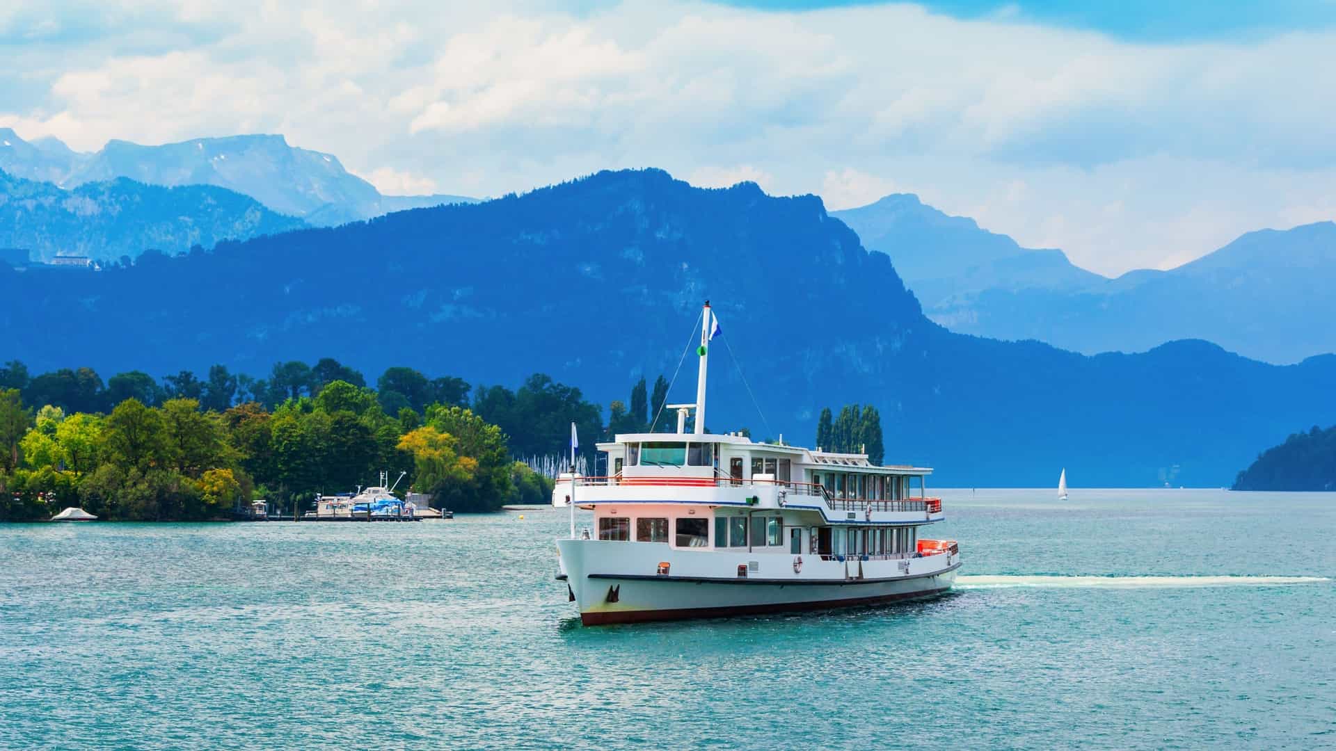 A scenic photograph of a tourist cruise boat sailing on the deep blue waters of Lake Lucerne in Switzerland. In the background, the cityscape of Lucerne is visible, with snow capped mountains and green hills rising in the distance under a clear sky.
