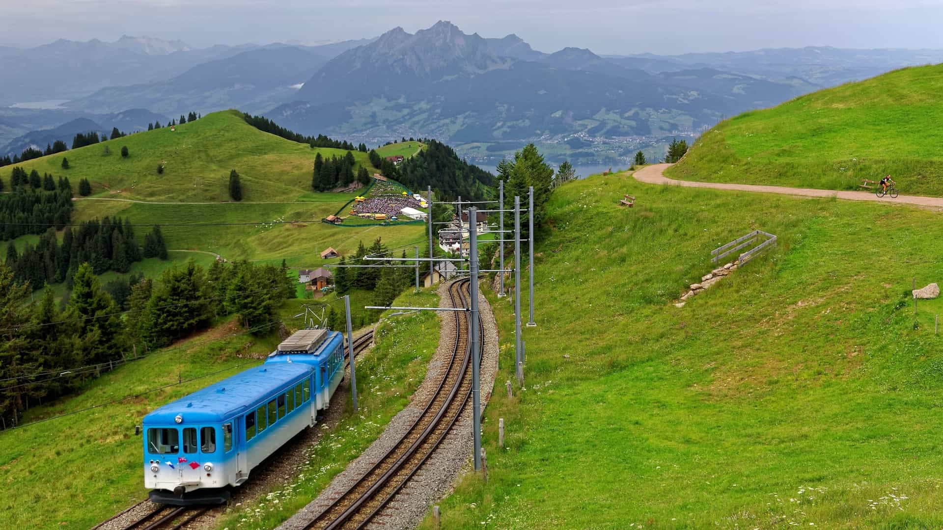 A scenic photograph of a red sightseeing train traveling on a cogwheel railway on Mt. Rigi in Switzerland. The train is moving through lush, green grassy meadows. In the distance, the rugged peaks of Mt. Pilatus and other majestic mountains are visible under a cloudy summer sky.