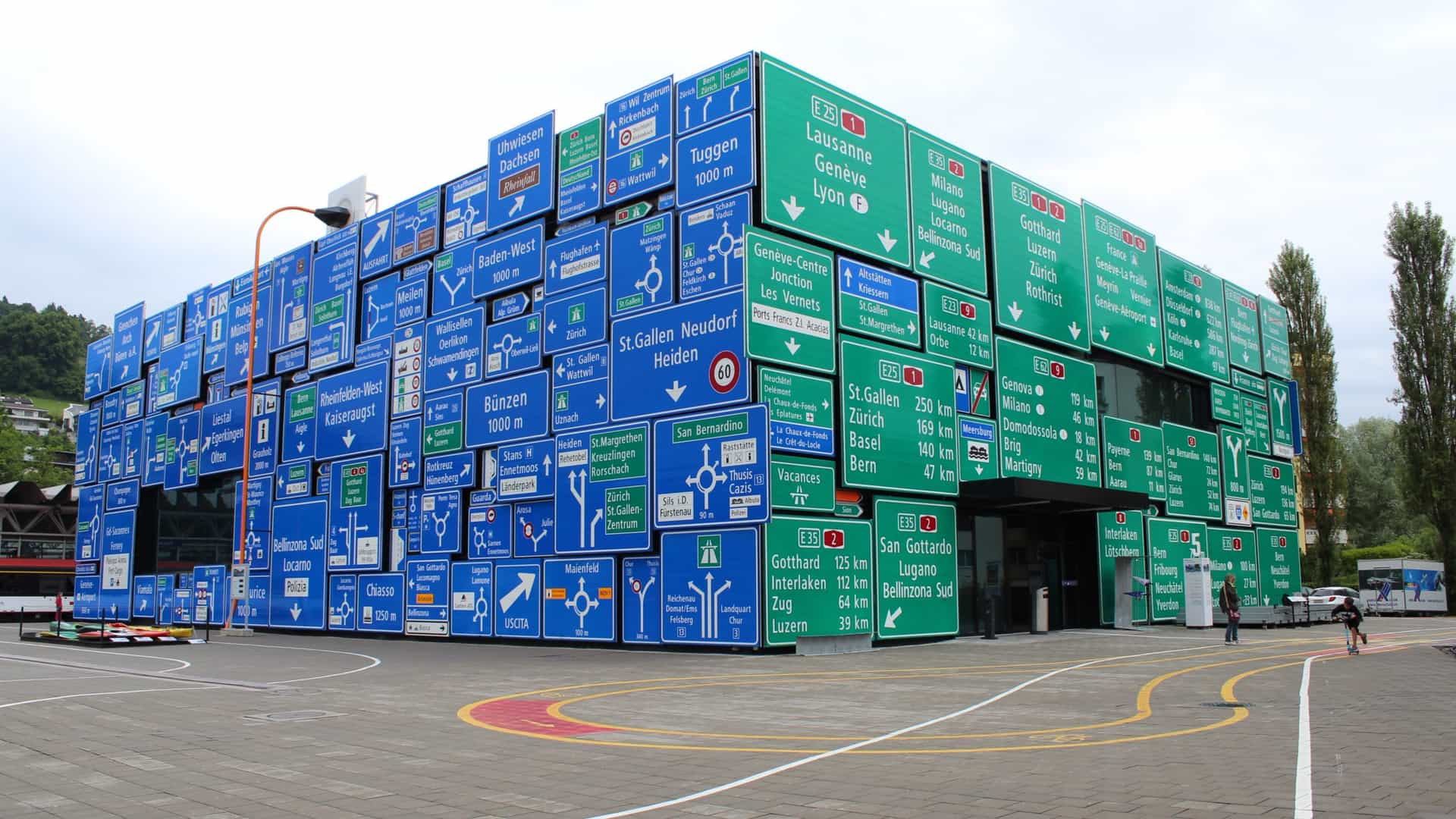 A detailed exterior view of the Road Transport Hall at the Swiss Museum of Transport (Verkehrshaus) in Lucerne, Switzerland. The building's facade is uniquely covered with a dense mosaic of hundreds of real Swiss road signs (such as warning signs and place-name signs), creating a recognizable and symbolic visual display for the transportation theme.