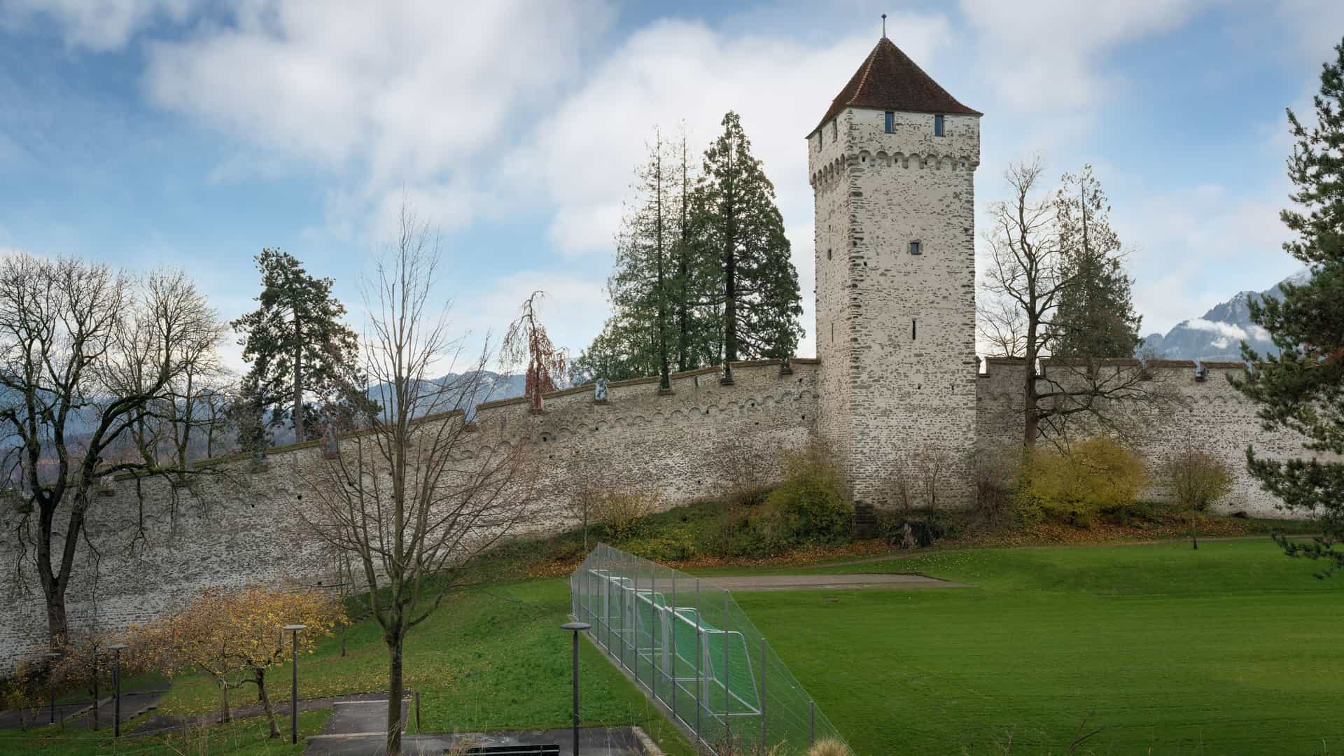 A photograph of the Zytturm (Clock Tower) in Lucerne, Switzerland, a medieval stone tower with a tiled roof that is part of the Musegg Wall. The tower's prominent feature is its large clock face on the facade, which is held by a painted mural of two giant figures. The clock has the unique privilege of chiming one minute before all other public clocks in the city.