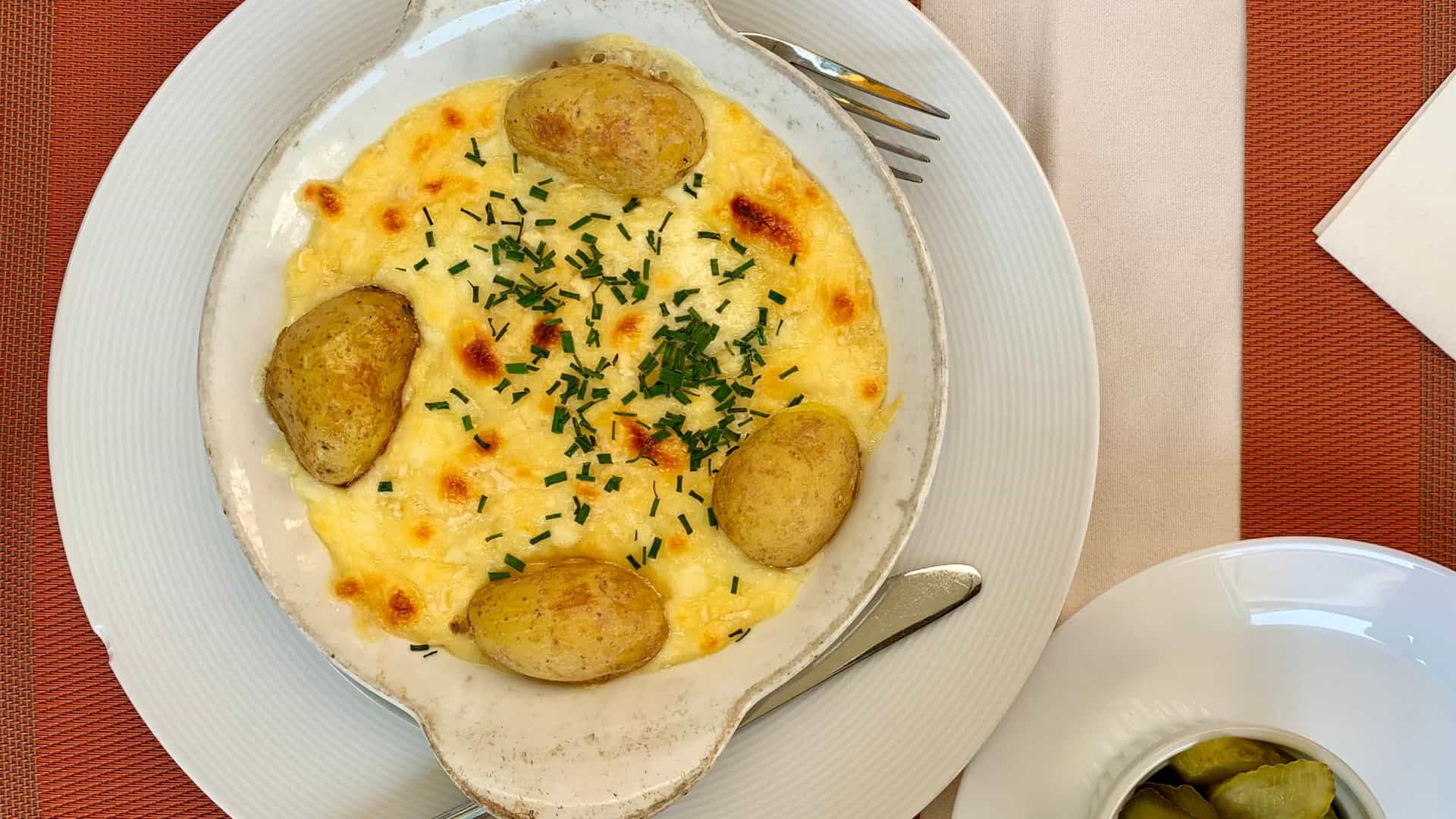 A close-up, top-down view of the traditional Swiss dish Raclette . The plate features melted Raclette cheese scraped over a serving of boiled potatoes, accompanied by small pickled gherkins and pickled onions on a rustic wooden table in a cafe setting.