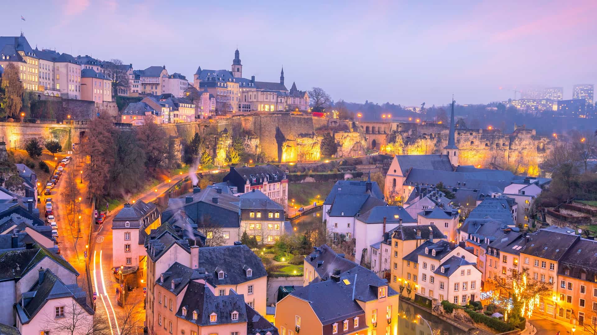 A top down view of the skyline of the old town of Luxembourg City. The image shows a dense cluster of historic buildings with red tiled roofs nestled within a valley, with the Alzette River winding through the scene. The perspective highlights the city's unique geography and dense, European style architecture.