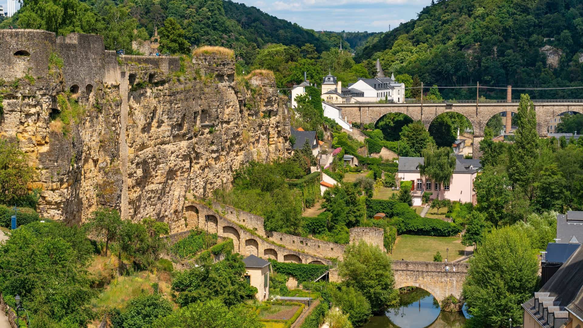 The Bock Casemates, a series of ancient tunnels and fortifications carved into a rocky cliff in Luxembourg City. The image shows the fortified walls and towers built into the rock, with the green valley and historic buildings of the Grund and Clausen districts visible below. The casemates are a prominent part of Luxembourg's UNESCO World Heritage Site fortifications.
