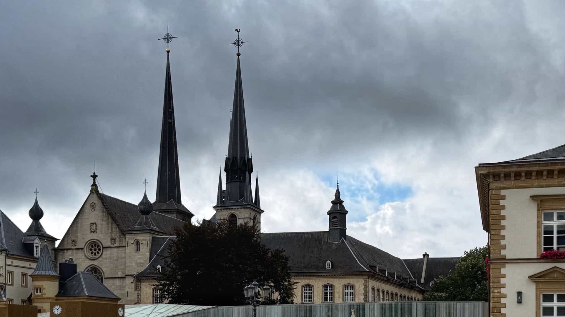 A view of the Notre-Dame Cathedral in Luxembourg City as seen from Place Guillaume II (Guillaume Square). The majestic Gothic and Renaissance spires of the cathedral rise above the historic buildings surrounding the square. The foreground often includes the Rodange memorial statue or other elements of the square.