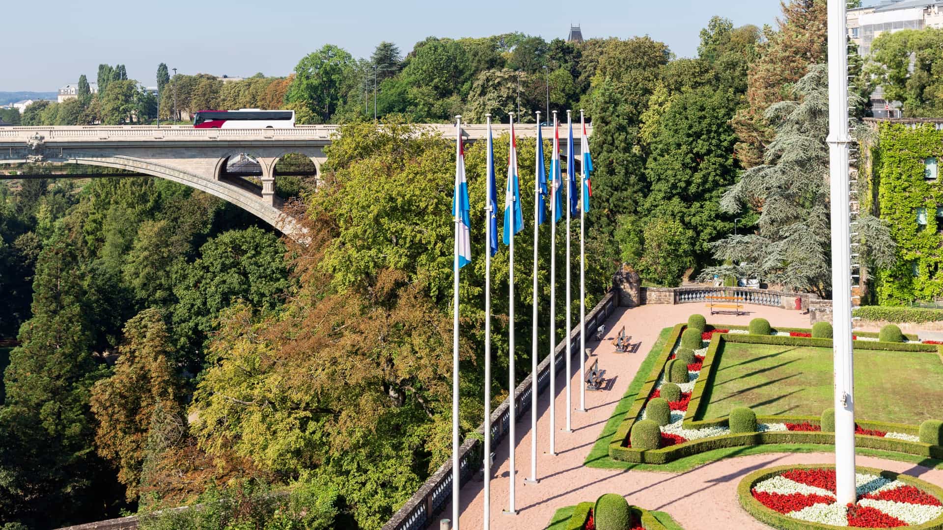 A scenic view of the Pont Adolphe (Adolphe Bridge) in Luxembourg City . The majestic stone arch bridge spans the green, forested Pétrusse Valley (Pétrusse Park), with a clock tower and other historic buildings visible on the city plateau in the background.