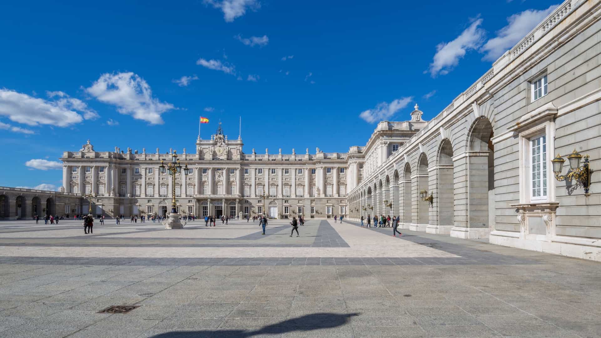 A view of the Royal Palace of Madrid on a bright, sunny day. The palace, a large and ornate white building with a classical facade, is set against a clear blue sky. In the foreground, there is a large plaza and green trees, with a fountain or monument in the center.