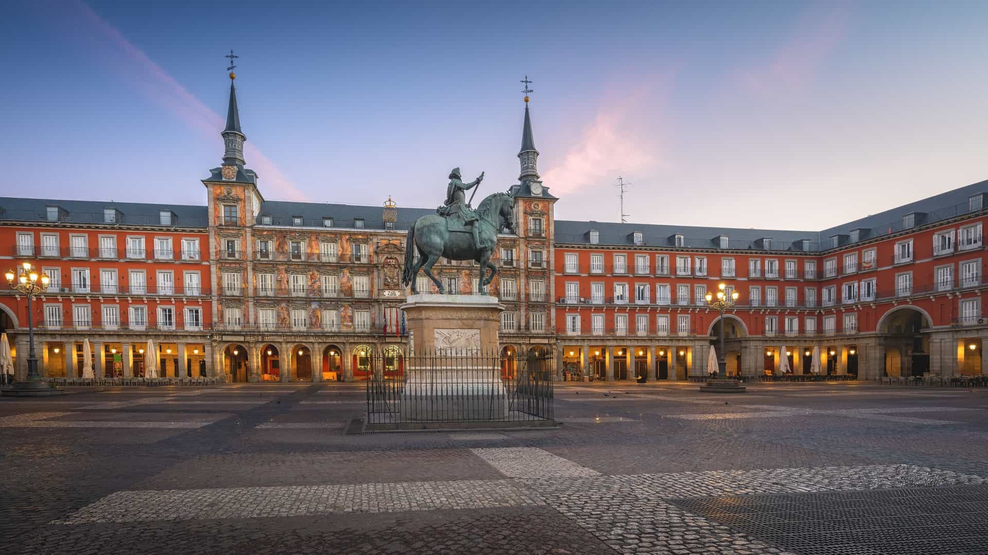 A view of the Plaza Mayor in Madrid at sunrise. The square is surrounded by multi story residential buildings with balconies and archways. In the center is the bronze equestrian statue of King Philip III on his horse, with a warm, golden light illuminating the scene.