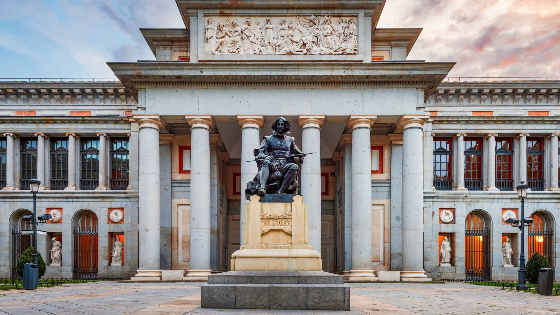A view of the exterior of the Prado Museum in Madrid, Spain. The building, a large, neoclassical structure with columns and multiple arched entrances, is situated on a wide, tree lined street. The building is a warm, reddish color, and there are people walking and standing in the foreground.