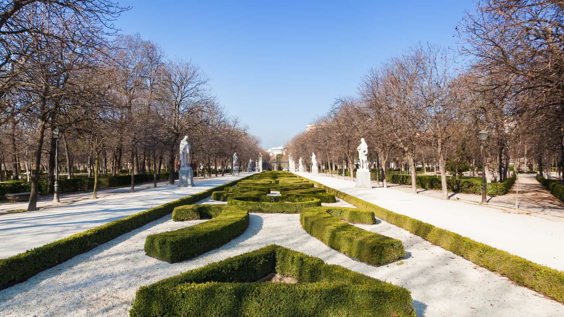 A wide angle view of the artificial lake in Retiro Park, Madrid, with the grand Monument to Alfonso XII visible in the background. The monument is a large, semicircular colonnade with an equestrian statue of the king at its center. Rowboats are visible on the lake, and the park's lush green trees line the shore.