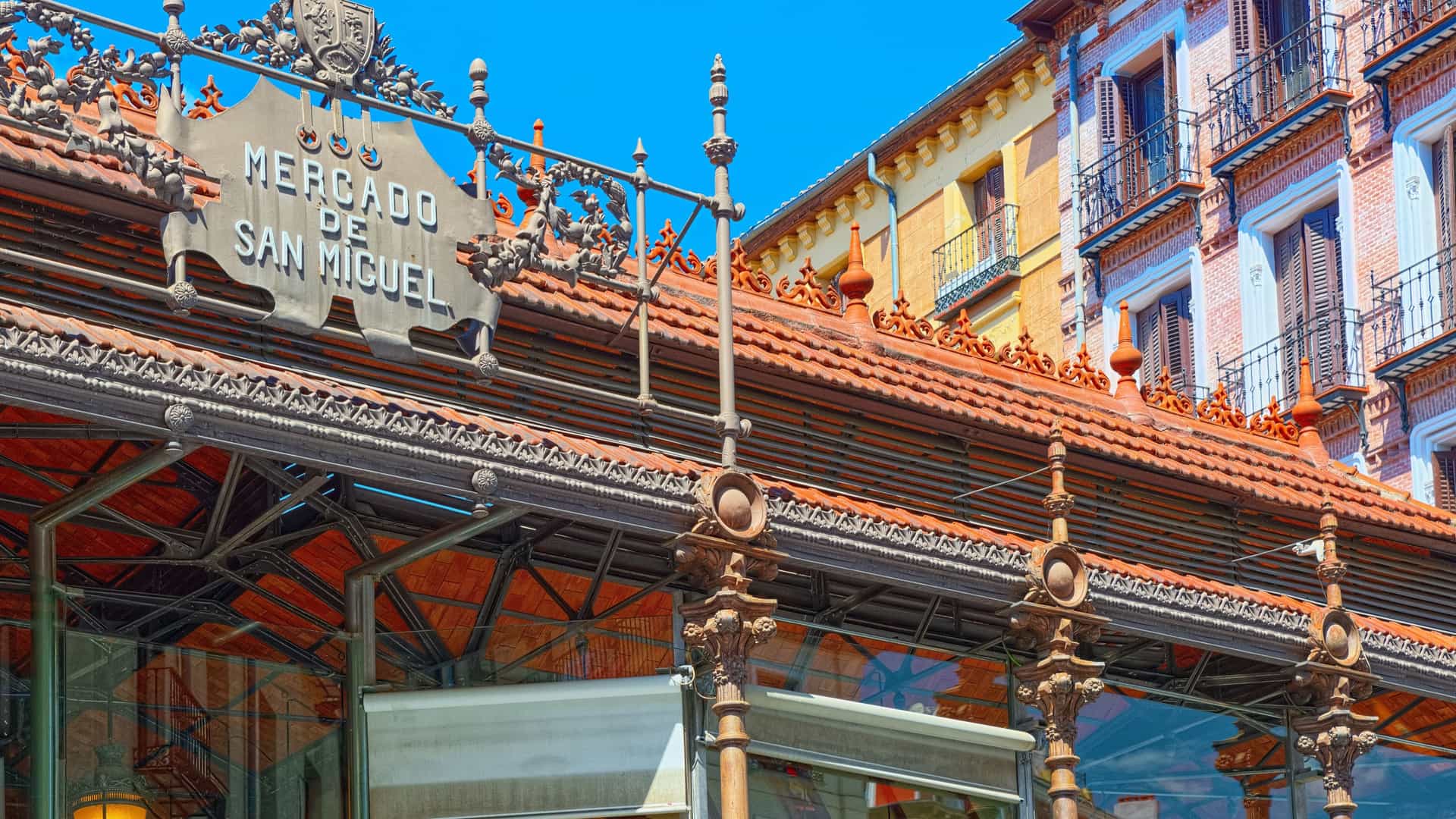 An interior view of the Mercado de San Miguel, a historic covered market in Madrid with a distinctive cast iron and glass structure. The market is bustling with people, and the stalls are filled with a wide variety of Spanish foods, including cured meats, cheeses, and freshly prepared tapas. The atmosphere is lively, with visitors standing at high tables and counters, eating and drinking.