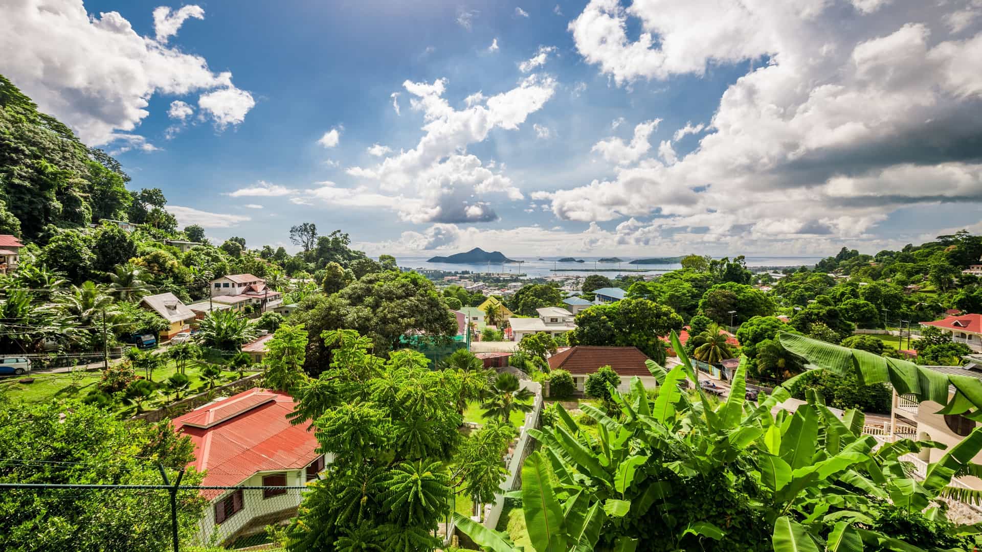 An elevated view of Victoria, the capital city of Seychelles, located on the northeastern coast of Mahé Island. The image shows the city nestled between a lush green, mountainous landscape and a clear blue sea, with a harbor filled with boats. Buildings with red rooftops are visible, and the overall scene depicts a tropical island capital.