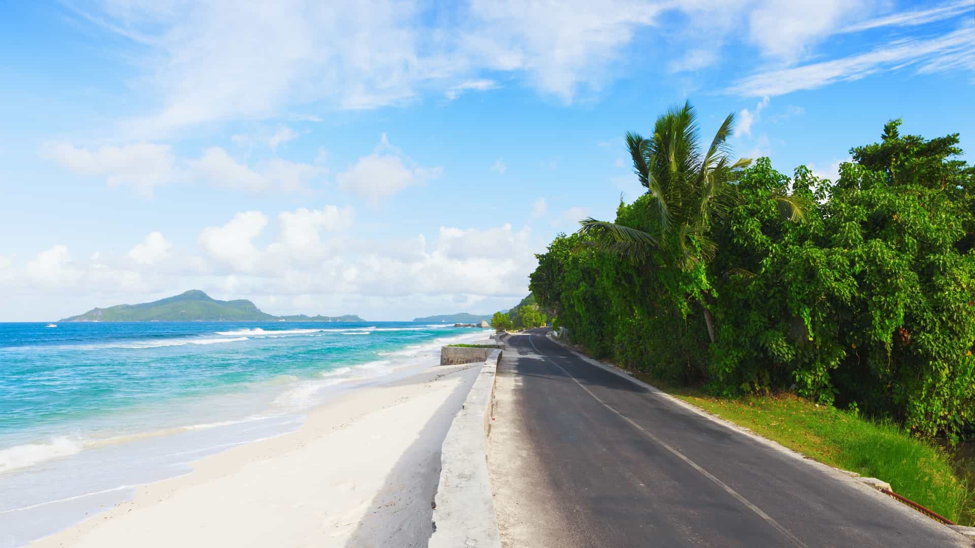 A beautiful tropical scene showing a narrow, paved Scenic Drive Along the Coastal Road running parallel to a stretch of white-sand beach in the Seychelles . The road is bordered by dense, lush green tropical foliage and palm trees on one side, and the brilliant turquoise water of the Indian Ocean and a barrier of seawall on the other. In the distance, a large, mountainous island is visible across the water under a blue sky with white clouds.