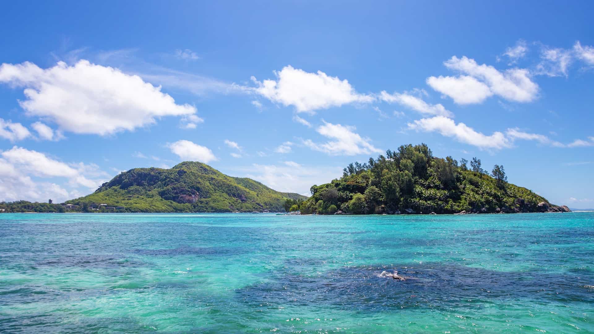 A lush, dome-shaped island covered in dense green tropical foliage, identified as Round Island in the Sainte Anne Marine National Park, Seychelles. The island is surrounded by the strikingly clear, turquoise and deep blue waters of the Indian Ocean. A small strip of white sand beach is visible along the shoreline, and the image captures the pristine natural beauty of this protected marine area.