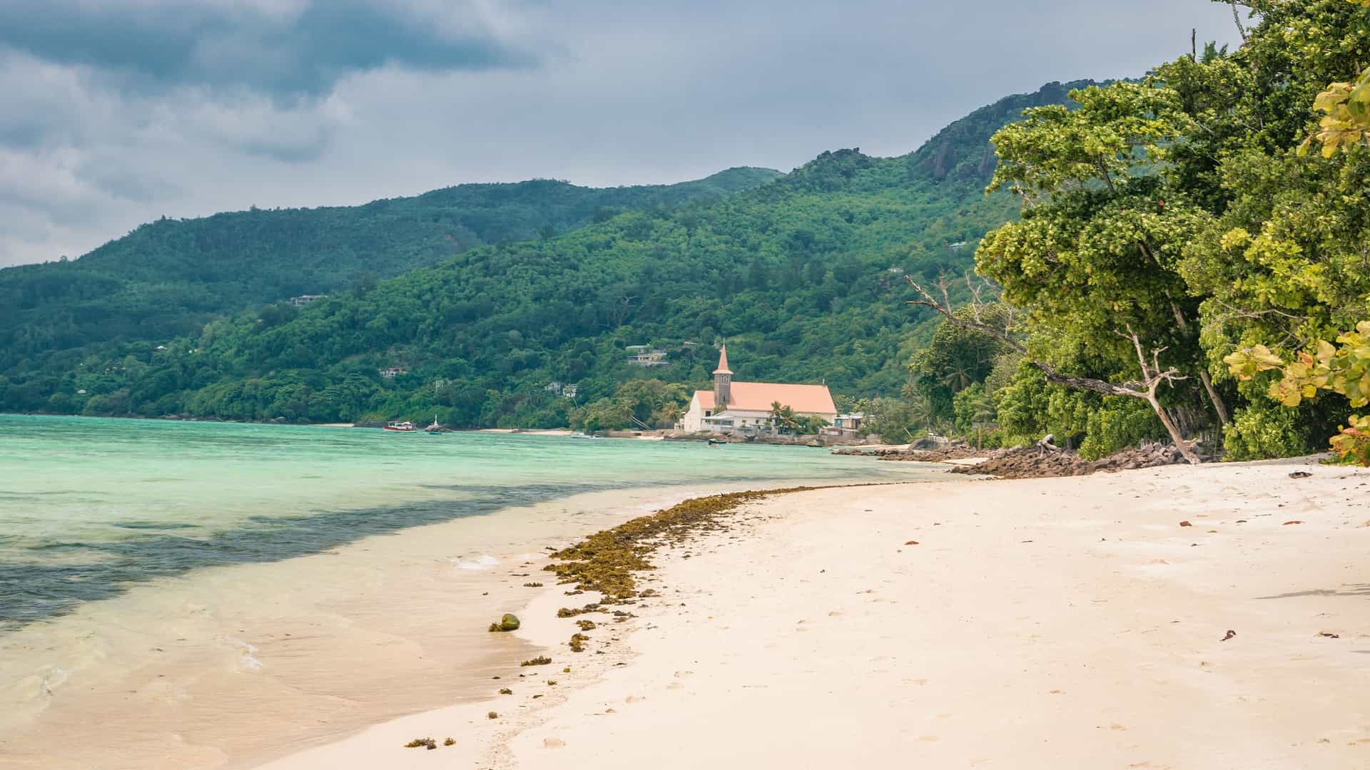 A picturesque view of the Saint Joseph, the Worker church situated on the coast of Anse Royale beach on Mahé Island in Seychelles. The image shows the church with a distinct steeple, a small cemetery, and lush green hills in the background, all overlooking the serene, clear turquoise waters of the Indian Ocean. The scene captures the unique blend of architecture and natural beauty.