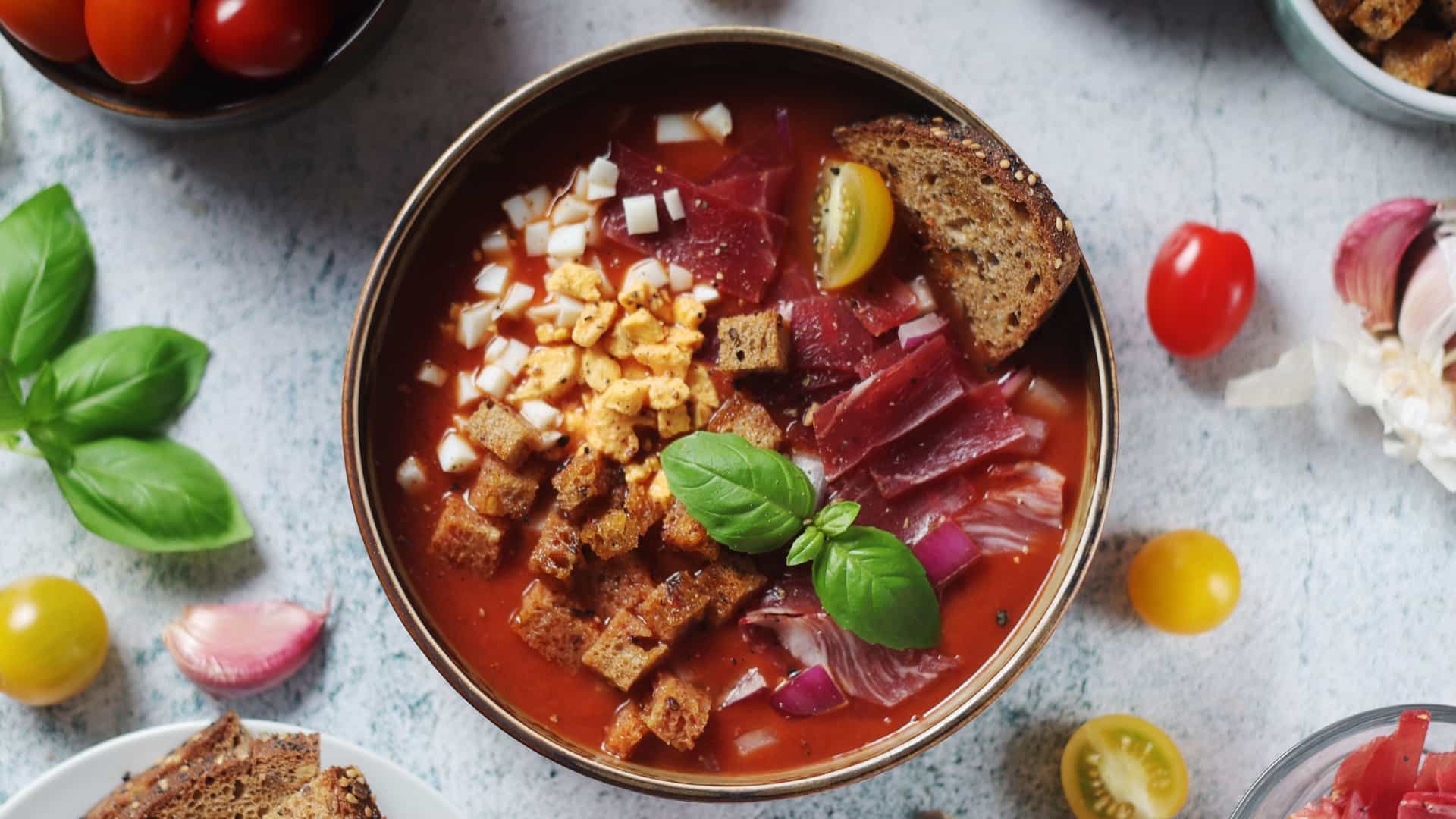 A vibrant bowl of salmorejo, a traditional Spanish cold tomato soup originating from Córdoba, Andalusia. The thick, creamy soup is topped with common garnishes, including finely chopped hard boiled egg, diced jamón serrano, and a drizzle of olive oil. The dish is presented on a rustic looking surface, highlighting its simple yet elegant presentation.