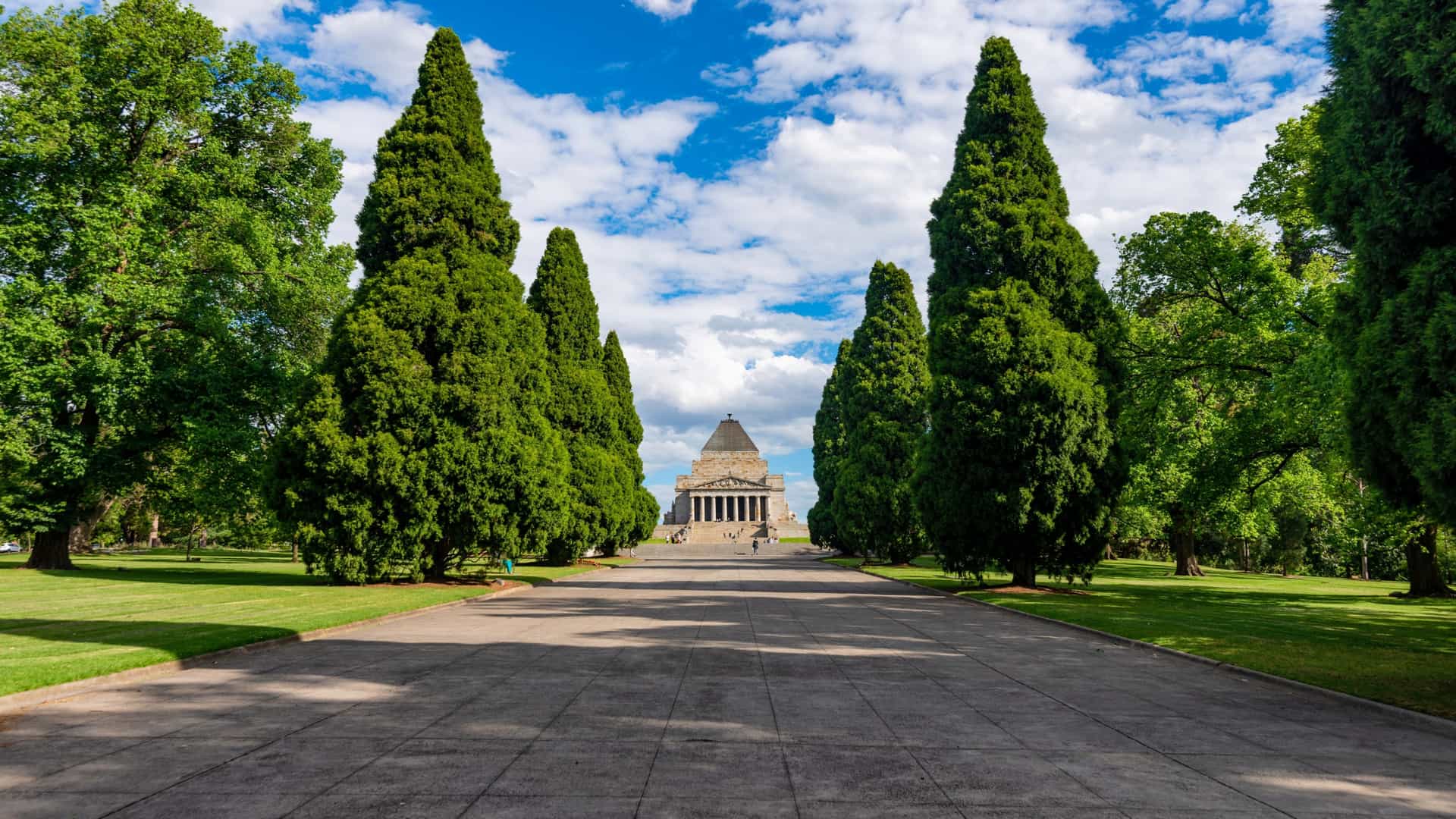 The Shrine of Remembrance in Melbourne, Australia, a solemn neoclassical war memorial. The image shows the impressive building, which is made of Tynong granite and features a large, pyramidal structure with prominent columns. It is surrounded by green lawns and trees, and the blue sky provides a serene backdrop, emphasizing its role as a place of reflection and commemoration for Australians who have served in conflicts.