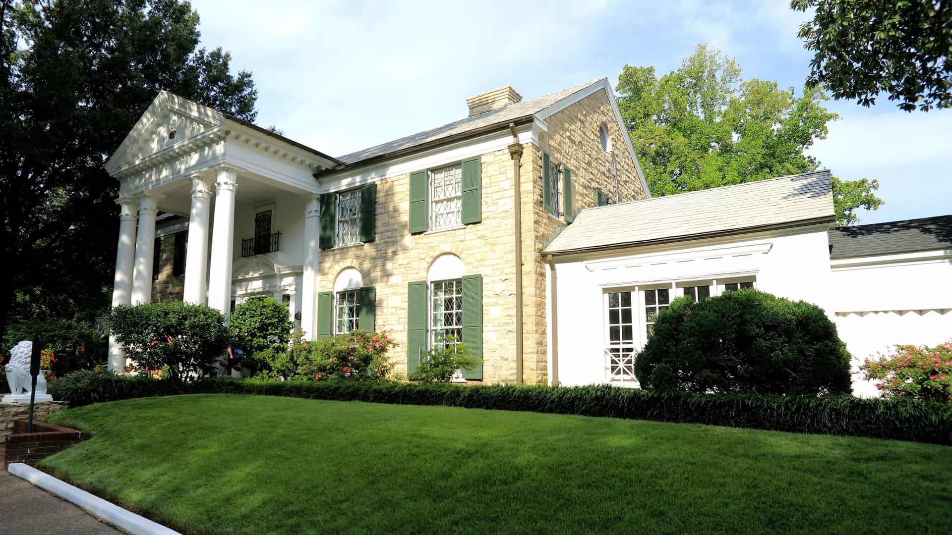 A stately, white-columned, neoclassical revival-style mansion, known as Graceland, is pictured in Memphis, Tennessee, USA. This iconic estate was the long-time home of the "King of Rock and Roll," Elvis Presley, and is now a U.S. National Historic Landmark surrounded by lush green grounds.