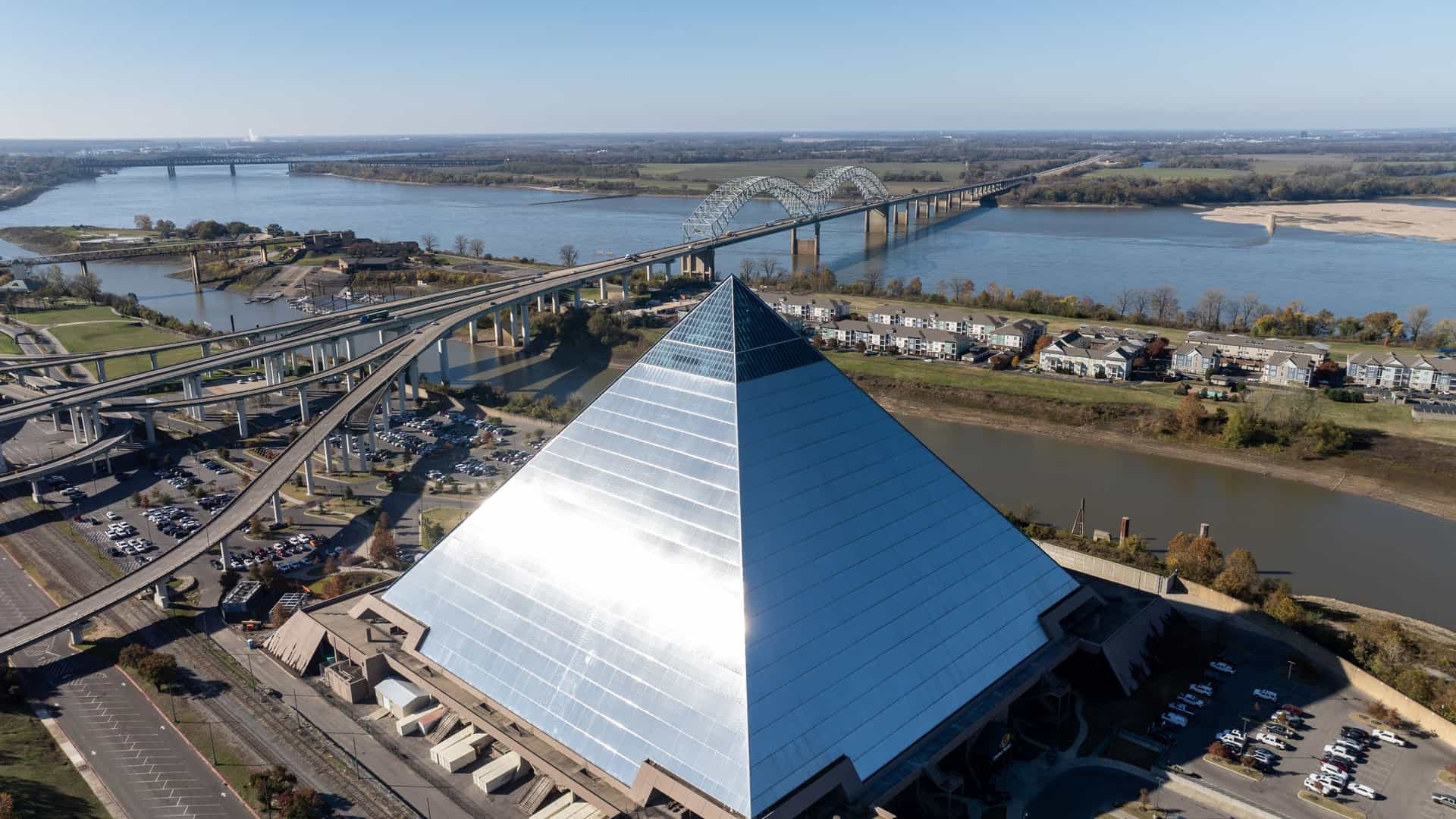 An aerial photograph featuring the massive, mirrored glass Memphis Pyramid structure in Tennessee, situated prominently near the banks of the Mississippi River. In the background, Interstate 40 traffic bridges, including the iconic Hernando de Soto Bridge, cross the wide river, framed by low-rise buildings and parking lots.