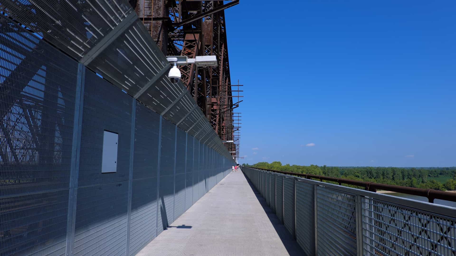 An expansive view of the Big River Crossing, the longest public pedestrian bridge across the Mississippi River, connecting Memphis, Tennessee, and West Memphis, Arkansas. The multi-use pathway is cantilevered off the side of the massive steel truss bridge (the Harahan Bridge), with the broad river and the city's waterfront visible under a bright sky.