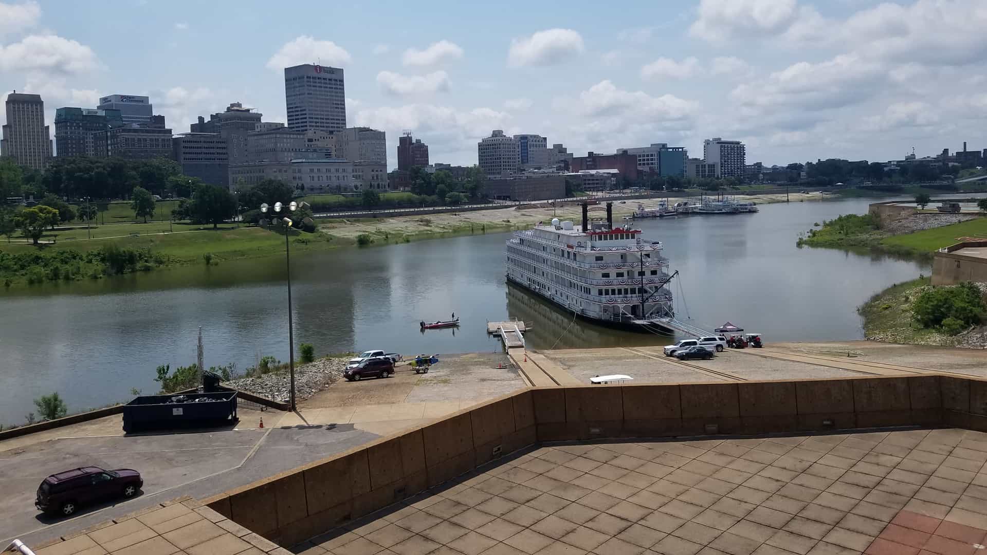 A wide-angle view of the downtown Memphis, Tennessee skyline in the fall, showing skyscrapers and a cruise on the Mississippi River. The Hernando de Soto Bridge is visible in the background, complemented by trees displaying beautiful autumn foliage.