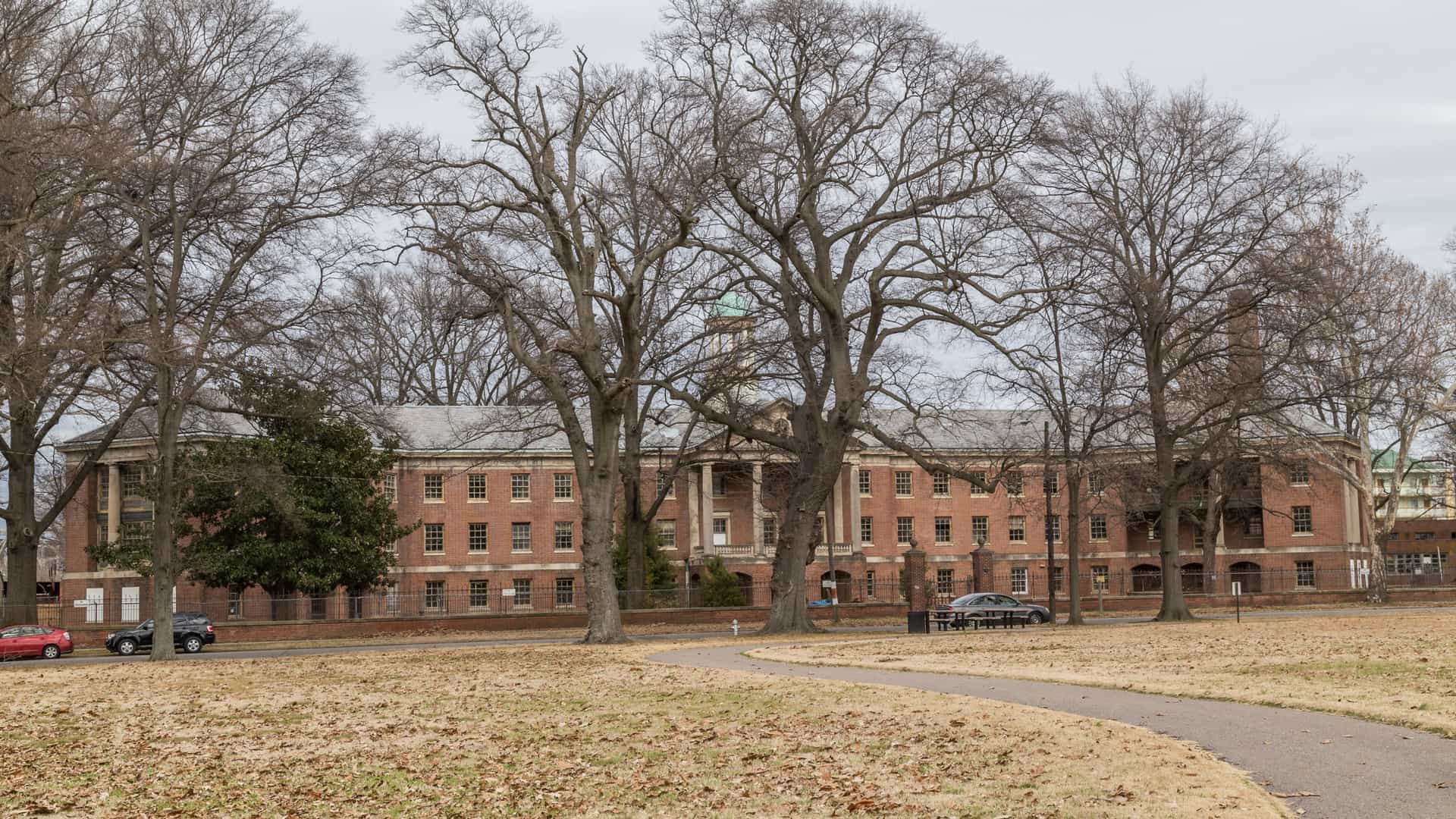 An exterior view of the historic Metal Museum in Memphis, Tennessee, featuring a grand, three-story red brick Italianate building with a central cupola. The museum is set on a lush bluff overlooking the Mississippi River, surrounded by manicured grounds and sculpture gardens under a clear sky.